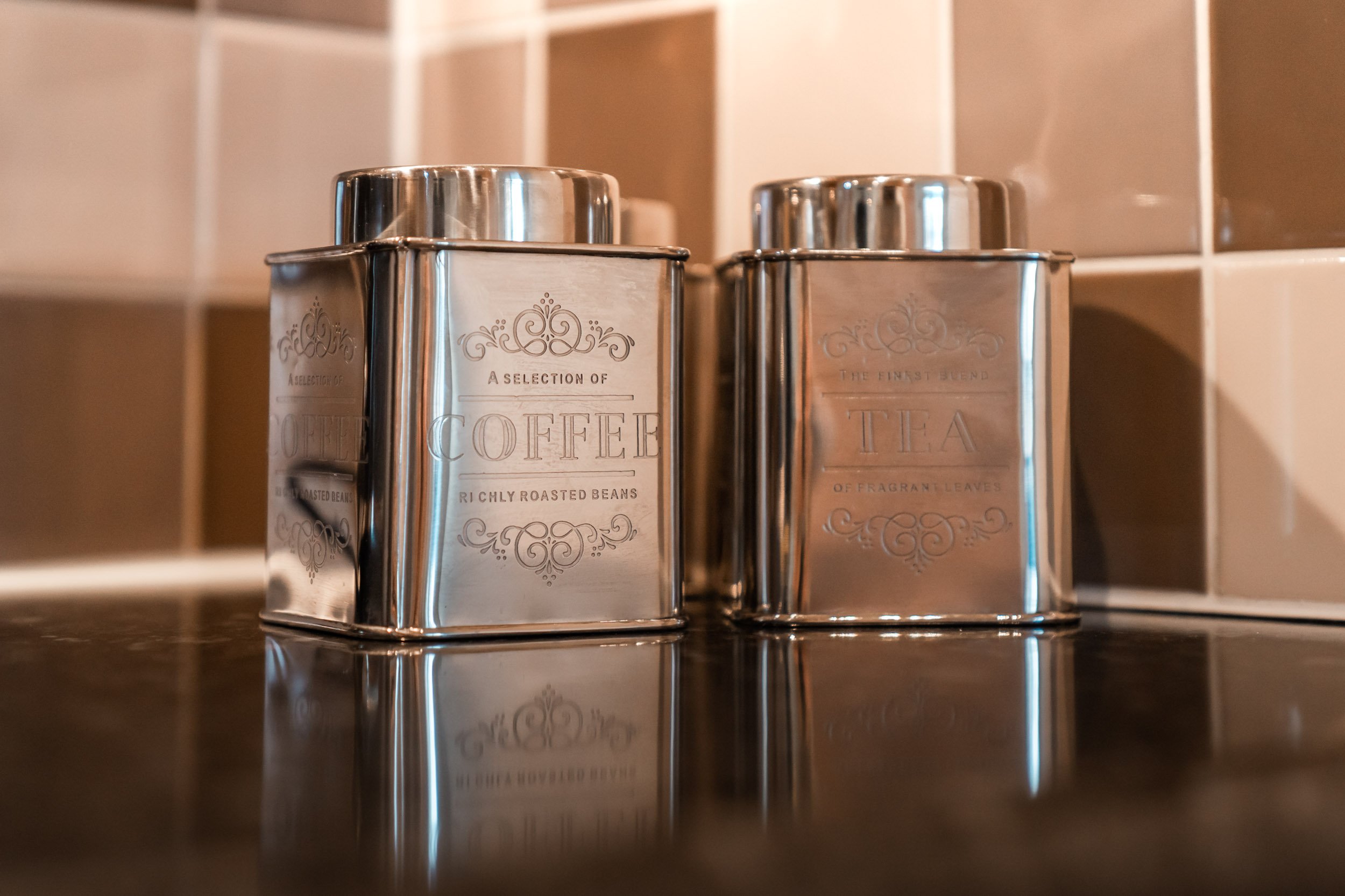 Two metallic containers labeled for coffee and tea on a kitchen counter with tiled backsplash.