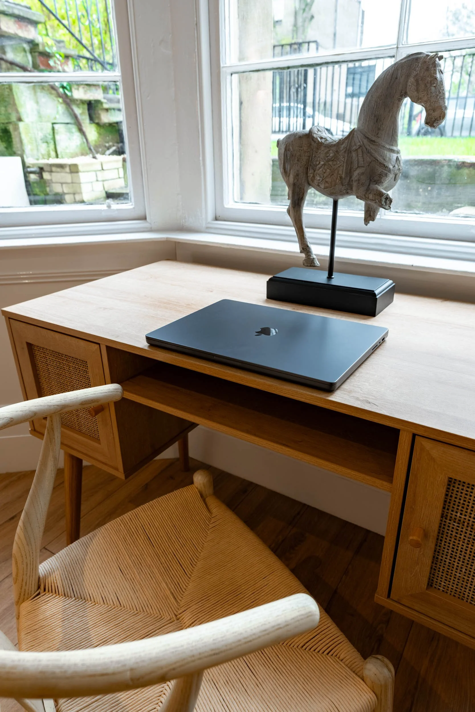 Wooden desk with a closed black laptop and a decorative wooden horse figurine on a stand, positioned in front of a large window with a view of a fenced garden area.