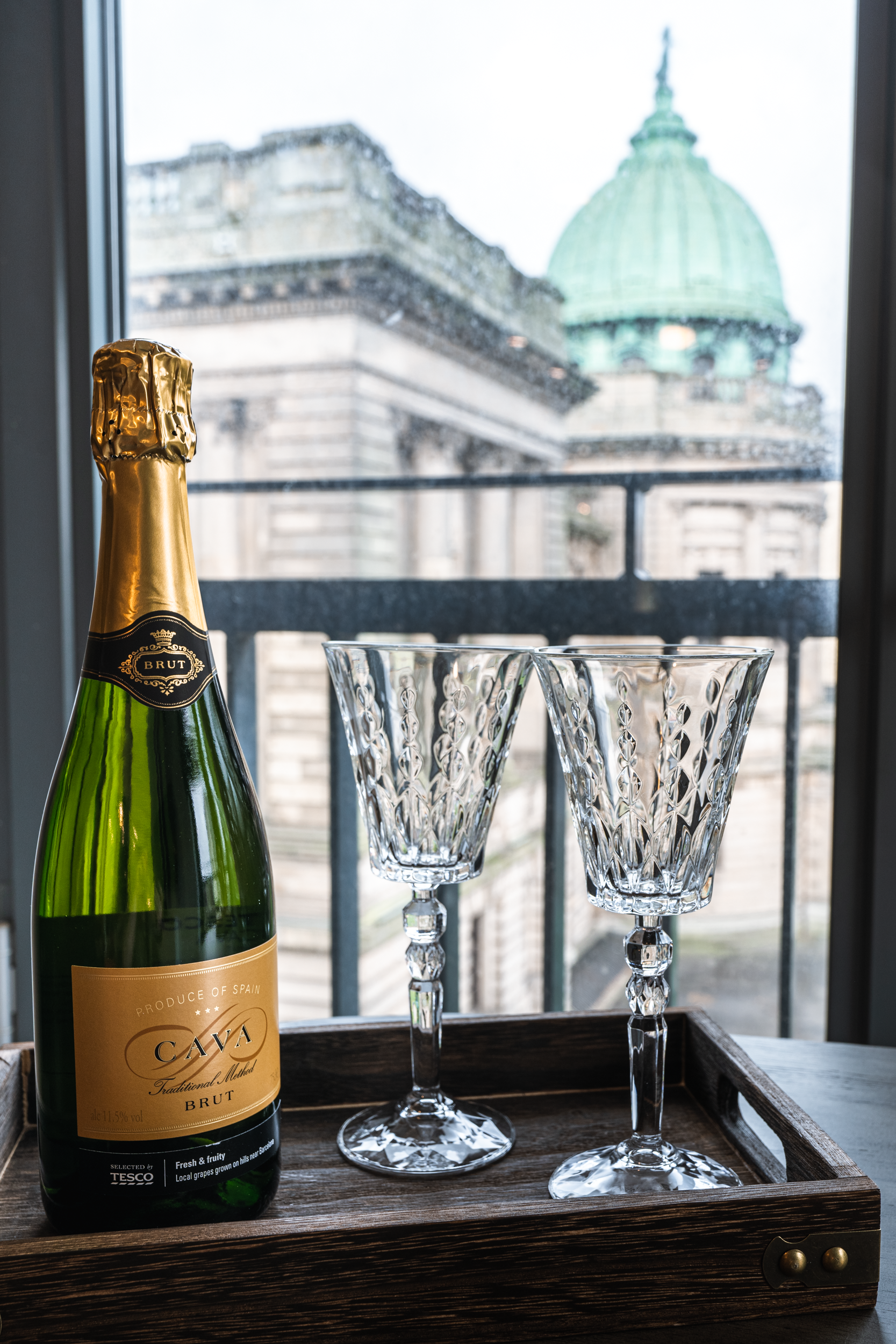 A bottle of sparkling wine and two empty crystal champagne flutes on a wooden tray, with a cityscape and a domed building visible through a window in the background.
