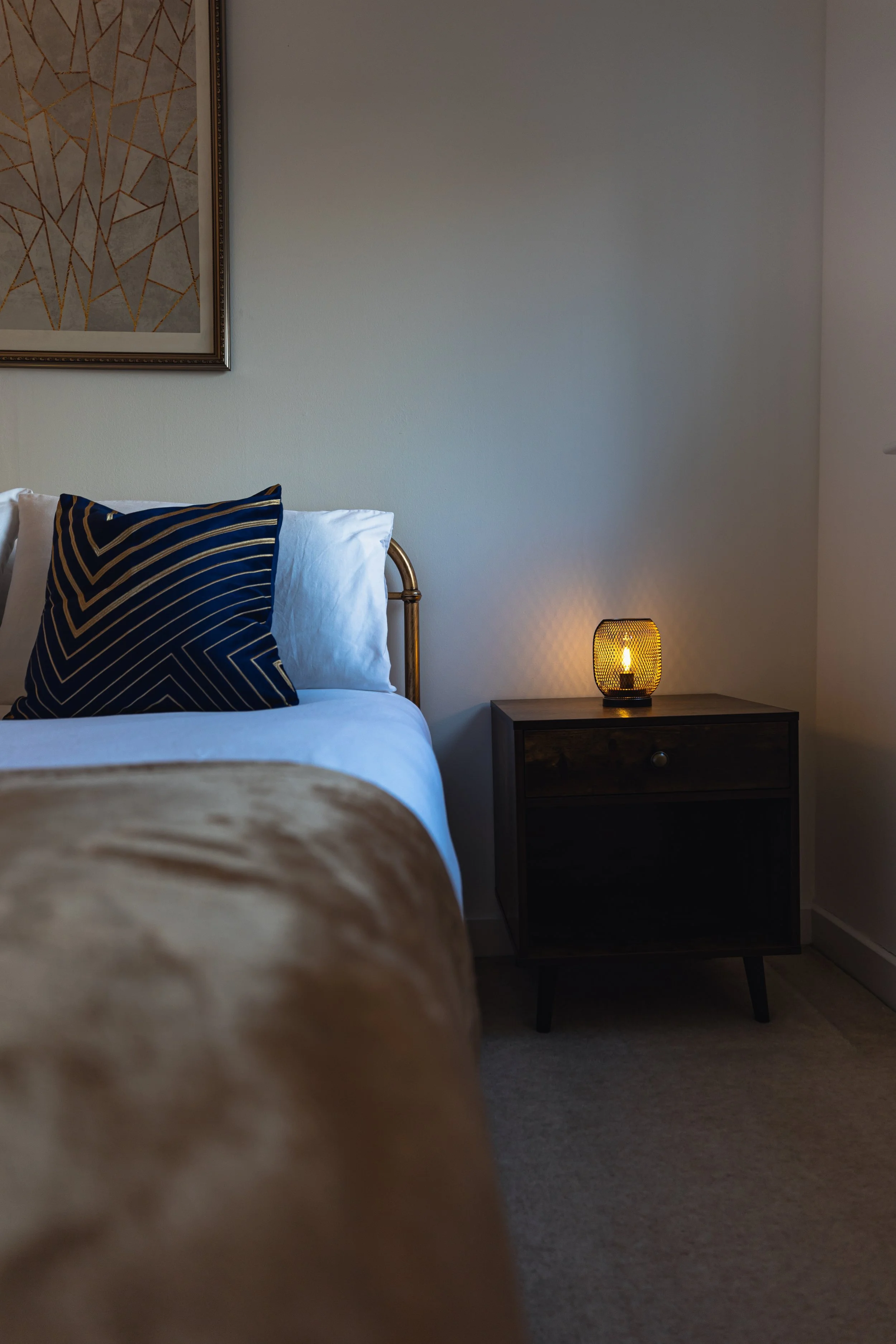 A bedroom corner with a bed featuring white sheets, a brown blanket, and a navy blue pillow with gold lines. A small dark wooden nightstand with a lit lamp is beside the bed, and a framed abstract artwork hangs above.