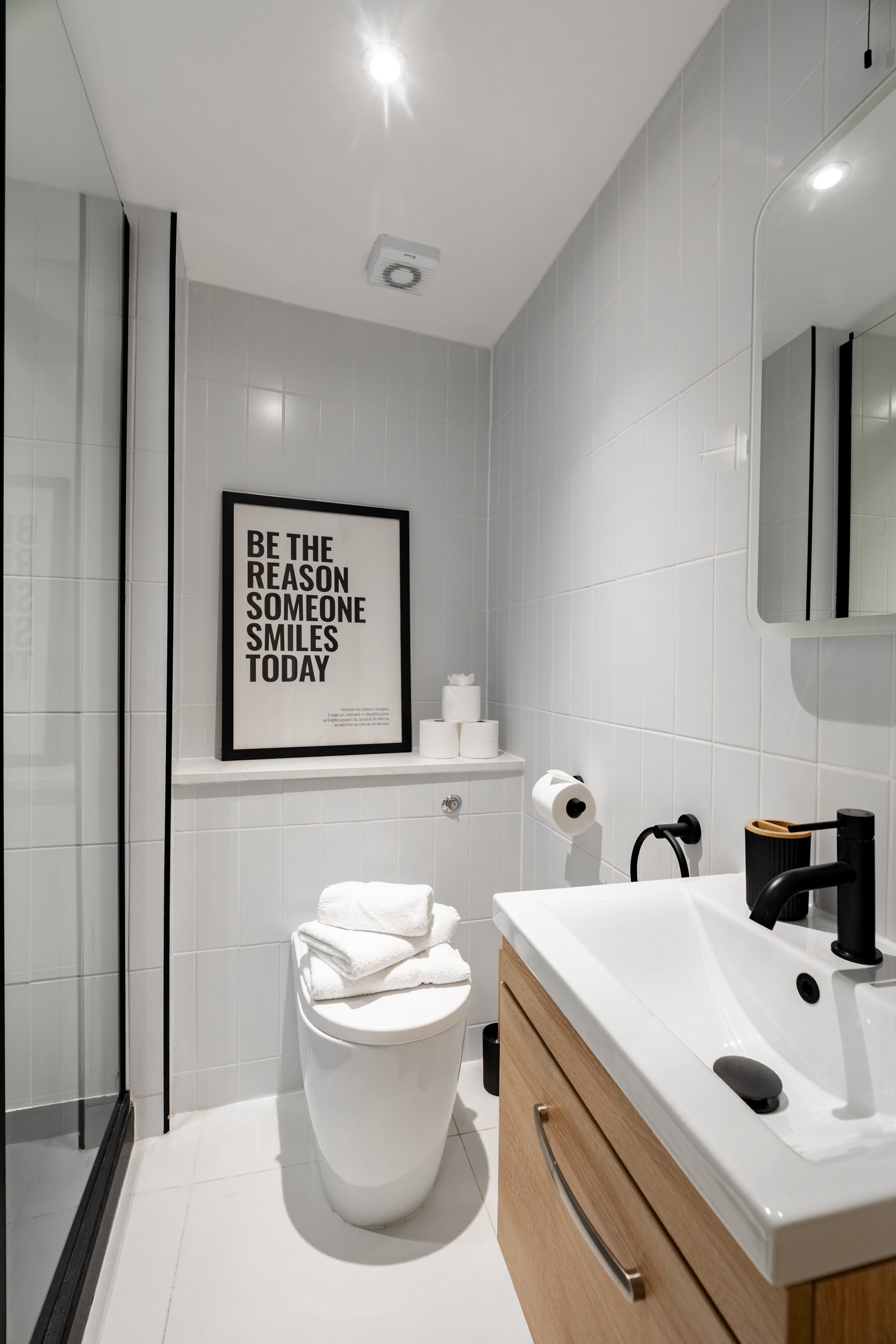 A modern bathroom with white tiles, black fixtures, a wooden vanity, a white toilet with towels on top, a framed motivational quote on the wall, and a small mirror.