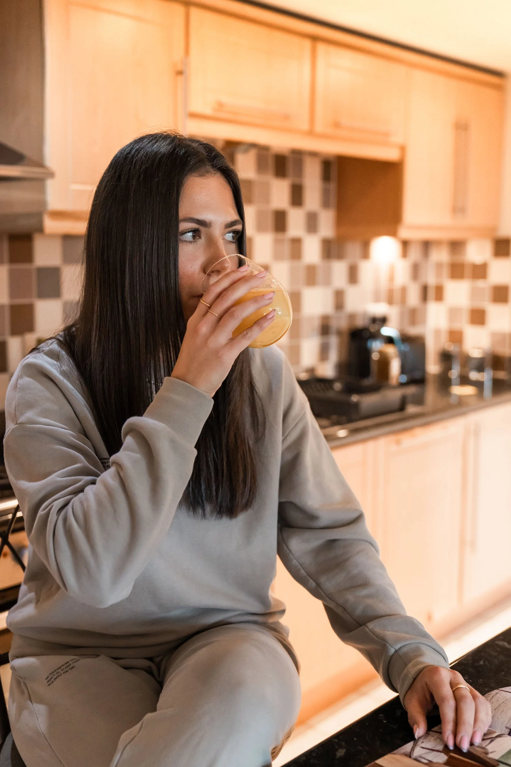 A woman with long dark hair sitting at a kitchen counter, drinking orange juice from a glass.