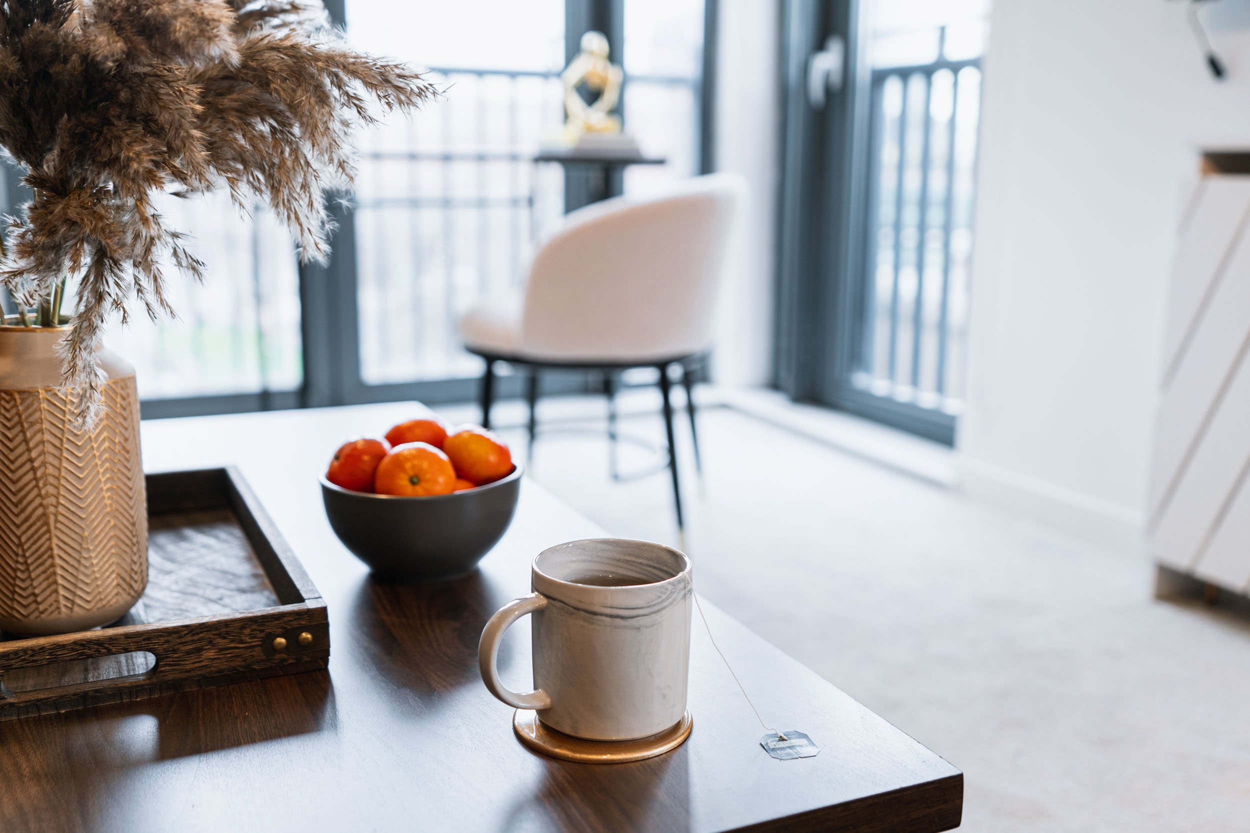 A kitchen table holding a potted plant, a black bowl with oranges, and a white mug with tea, in a room with large windows and a chair in the background.