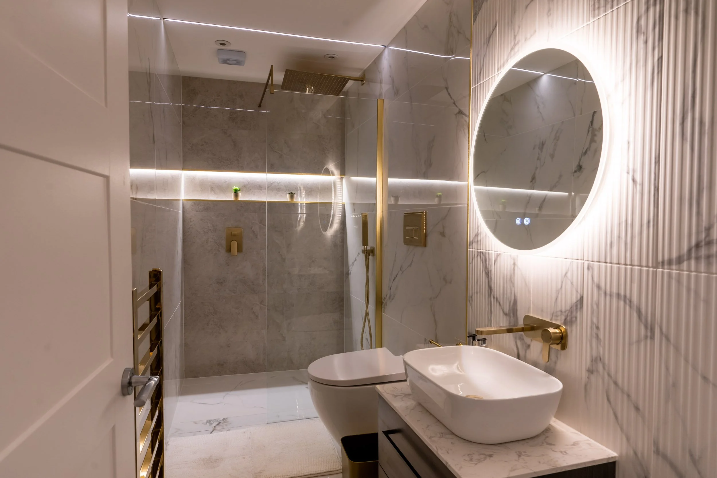 A modern, luxury bathroom with marble and textured wall tiles, illuminated mirror, vessel sink with gold fixtures, and a walk-in shower with a rainfall showerhead.