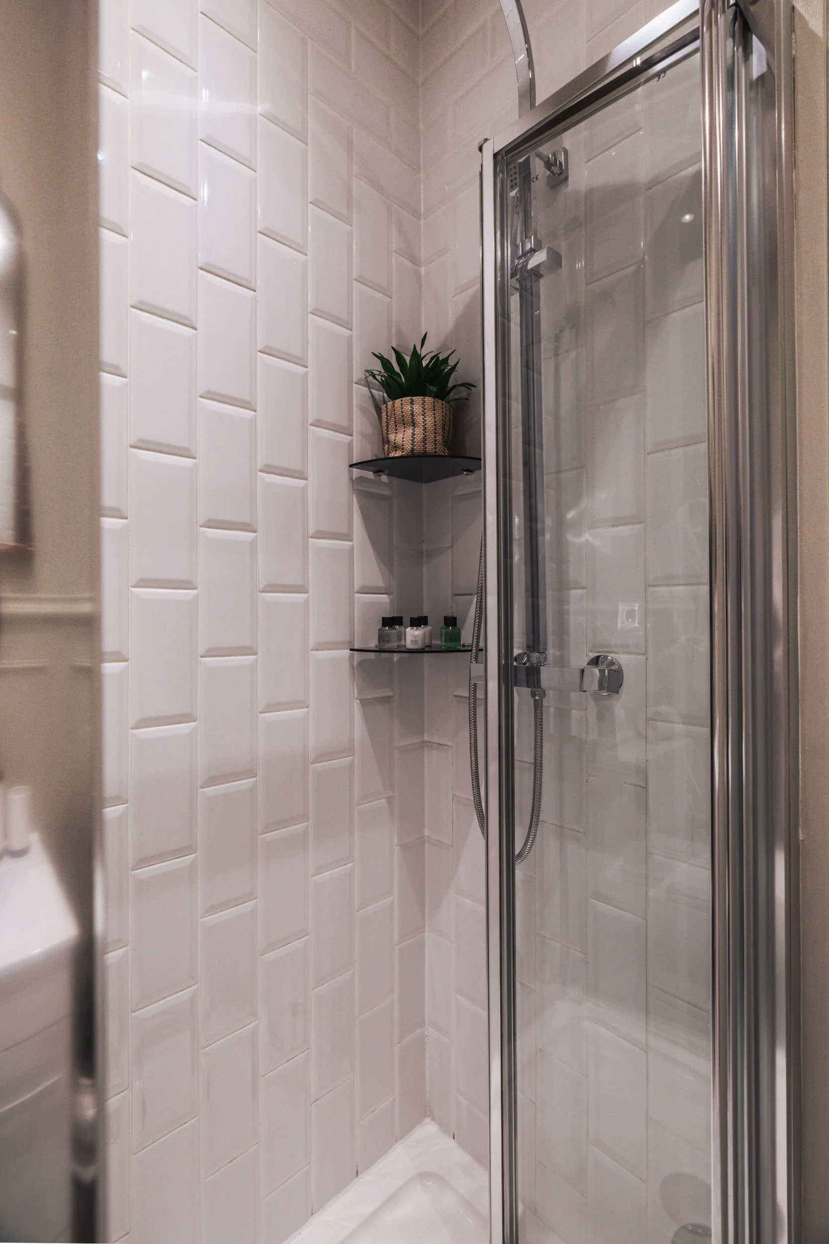 A small enclosed shower with white tiles on the walls, a sliding glass door, a handheld showerhead, a corner shelf with a plant in a woven basket, and several small bottles of toiletries.