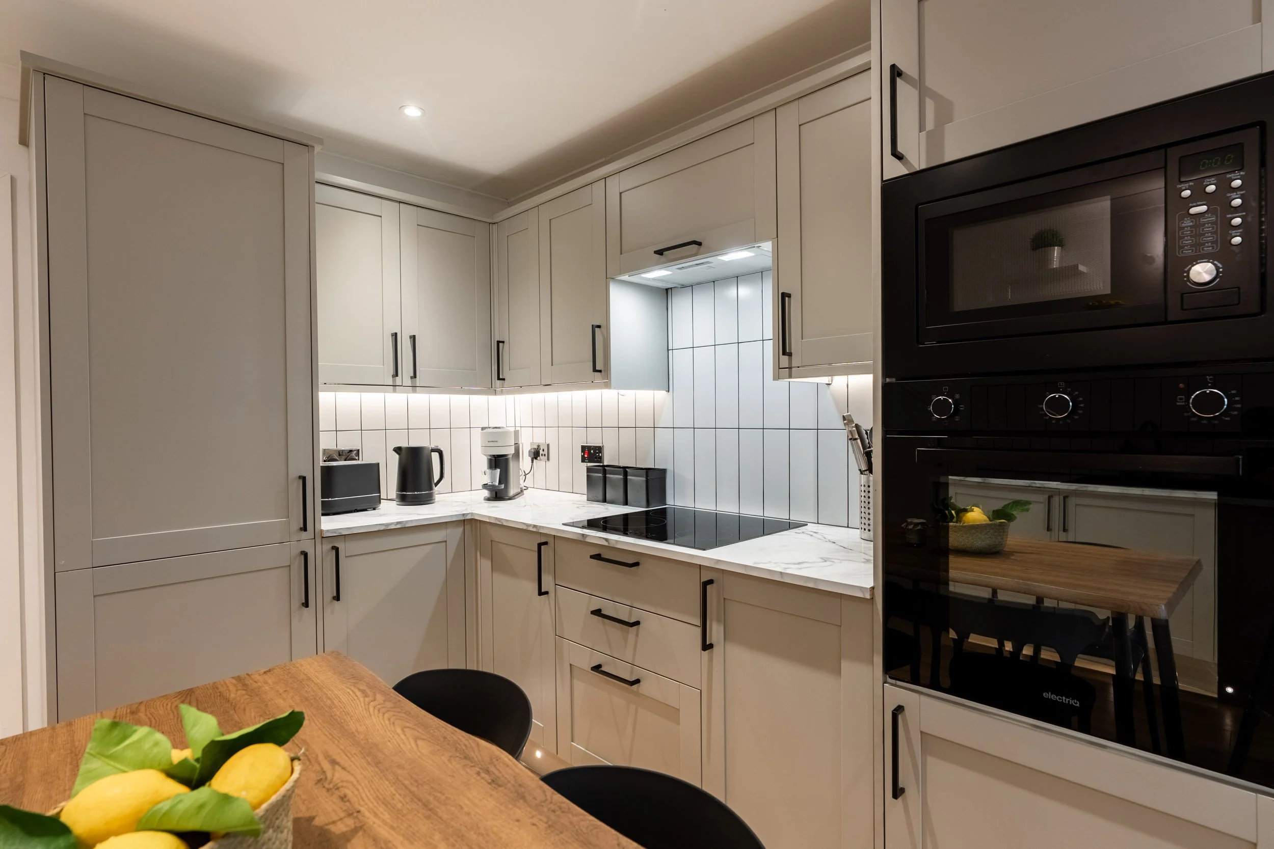 Modern kitchen with white cabinets, marble countertop, and black appliances, including microwave and oven, with a small dining table and lemons in a basket.