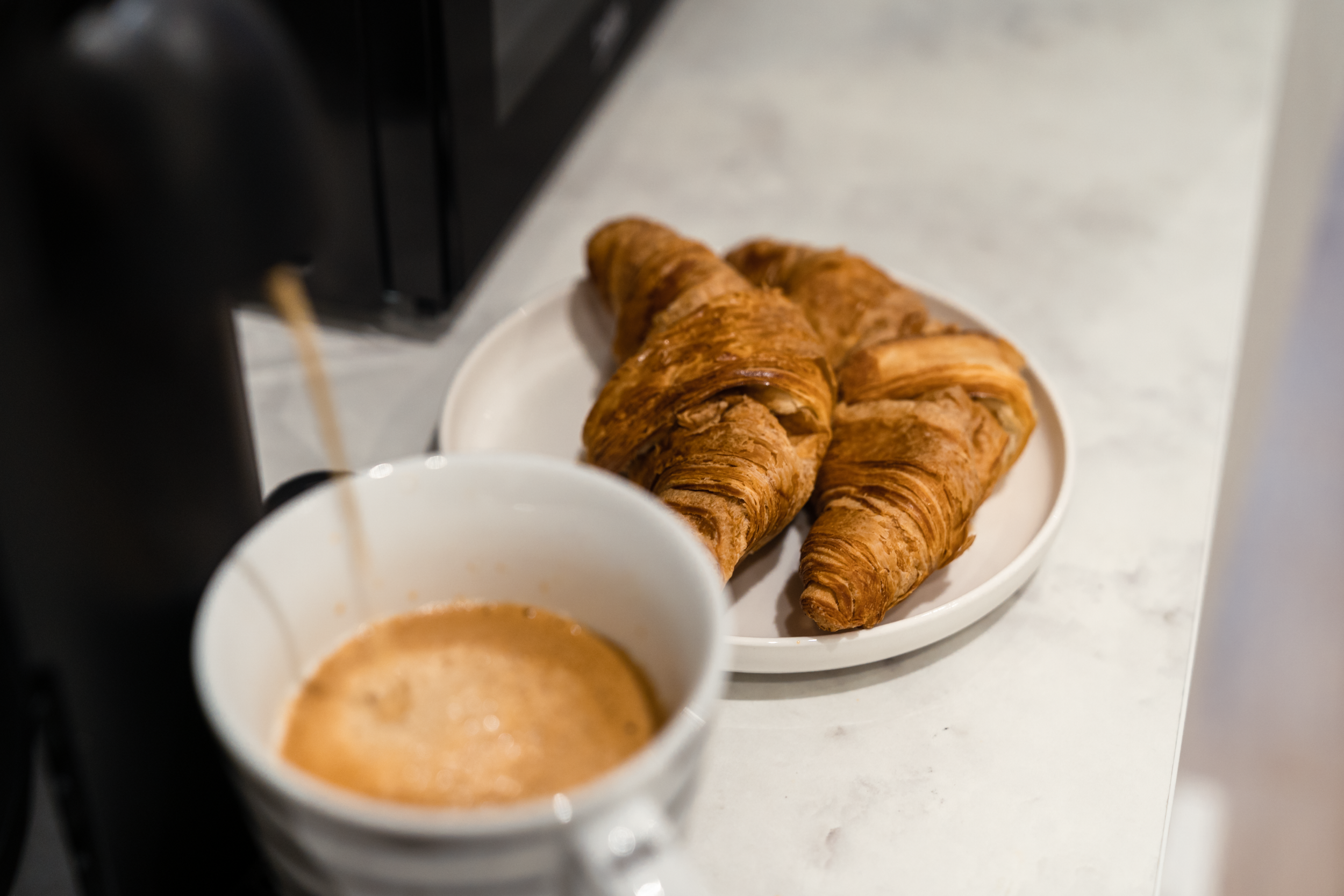Freshly brewed coffee being poured from a coffee machine into a white mug, placed on a white countertop with a white plate holding three golden brown croissants.