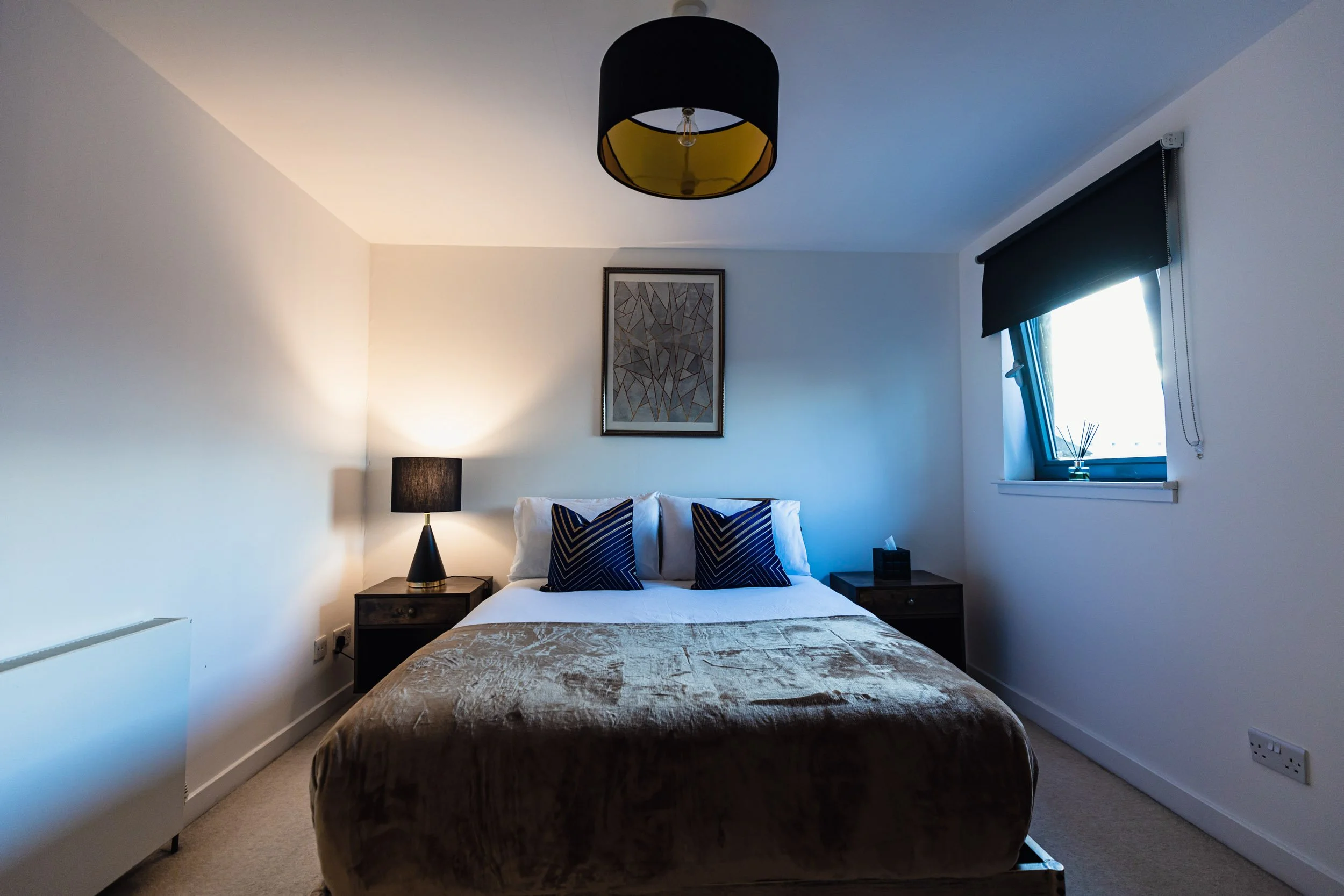 A bedroom with a bed, two bedside tables with lamps, a framed picture on the wall, a window with black blinds, and a ceiling light fixture.