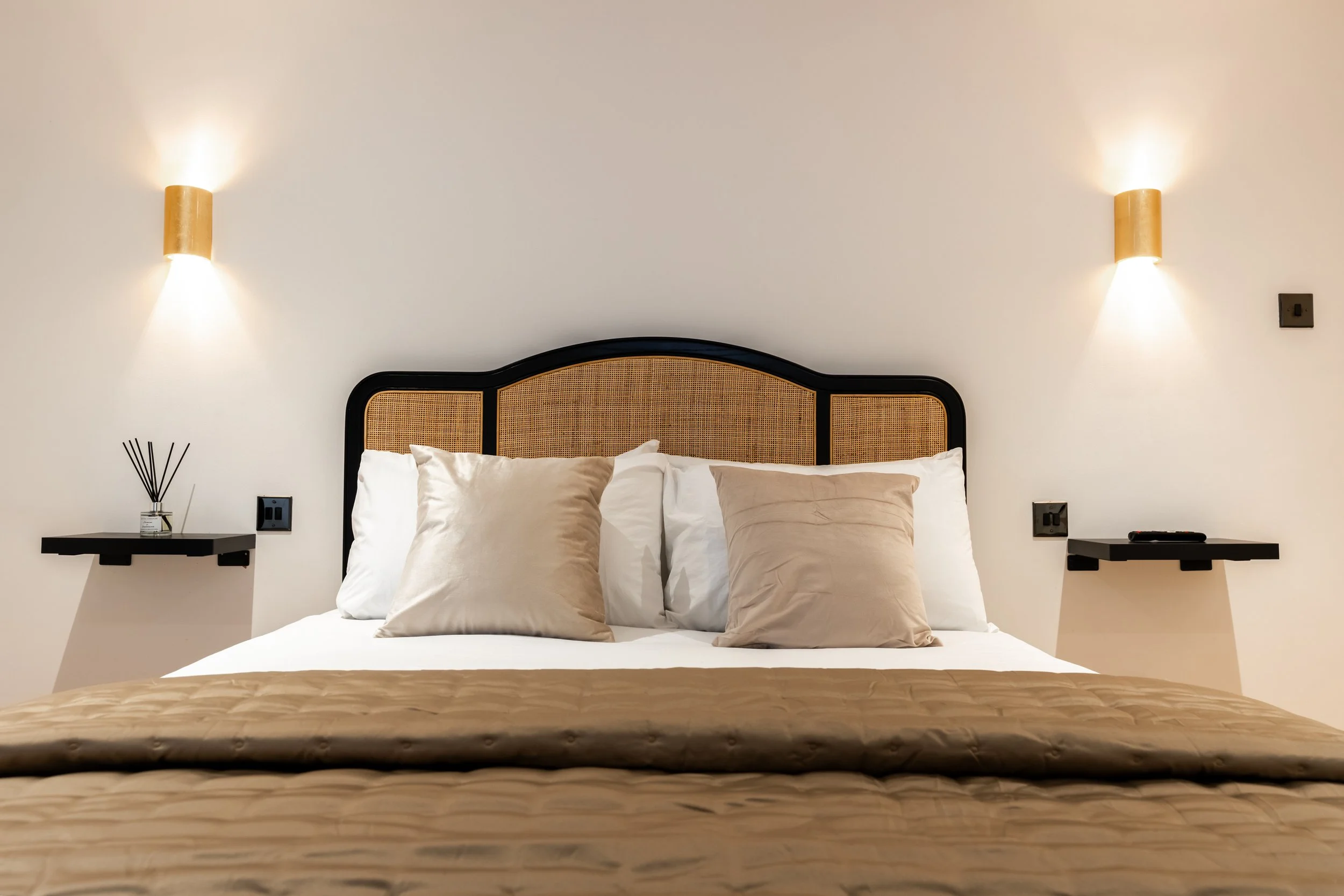 A neatly made bed with a black frame and rattan headboard, topped with beige and white pillows, beige bedding, and a beige bedspread. There are two wall-mounted lamps above each side of the bed and small black floating nightstands, one with a diffuse