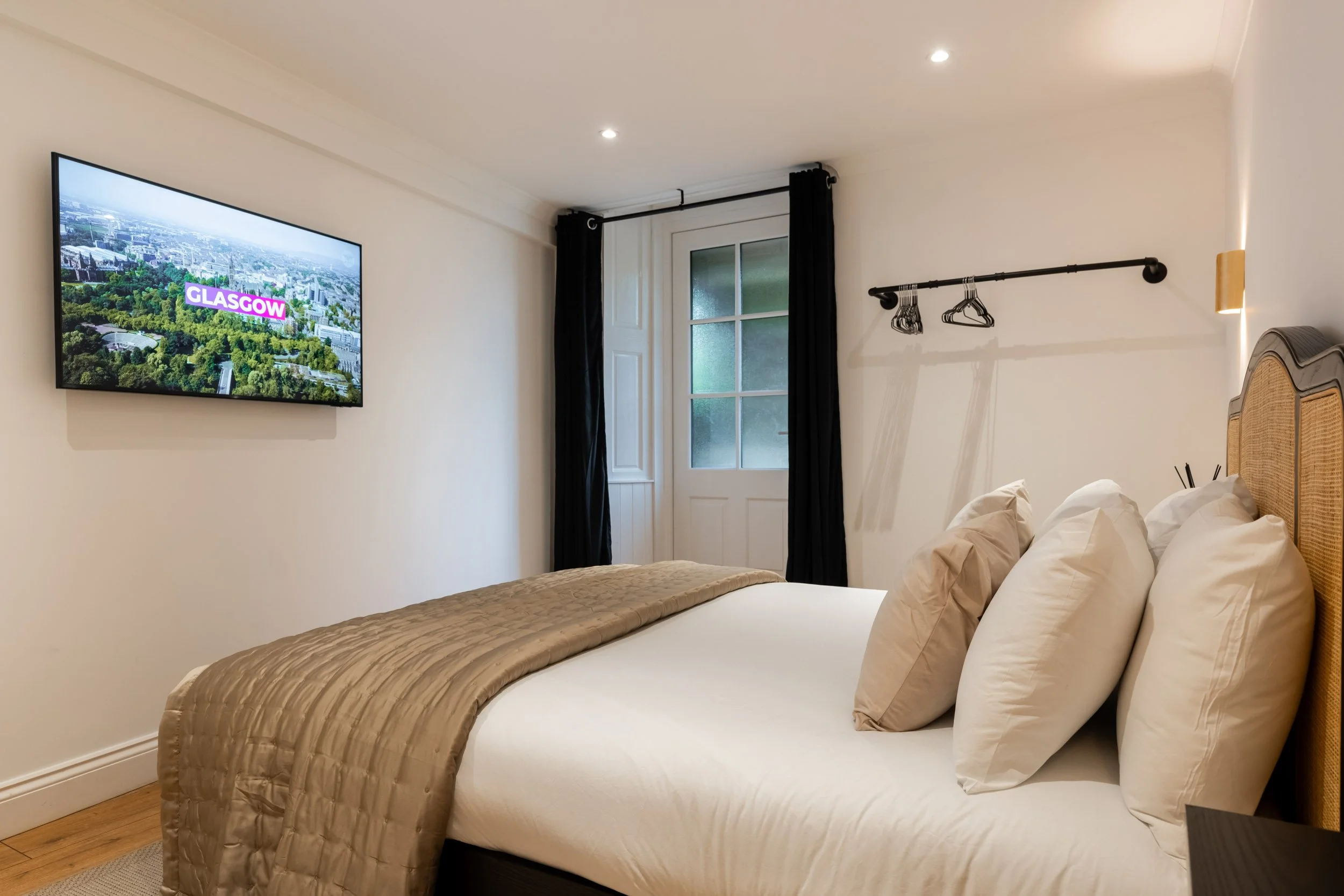 A simple hotel room with a bed, white bedding, and pillows, a wall-mounted television showing an aerial view of Glasgow, a door with frosted glass, black curtains, a closet rod with hangers, and a wooden headboard.