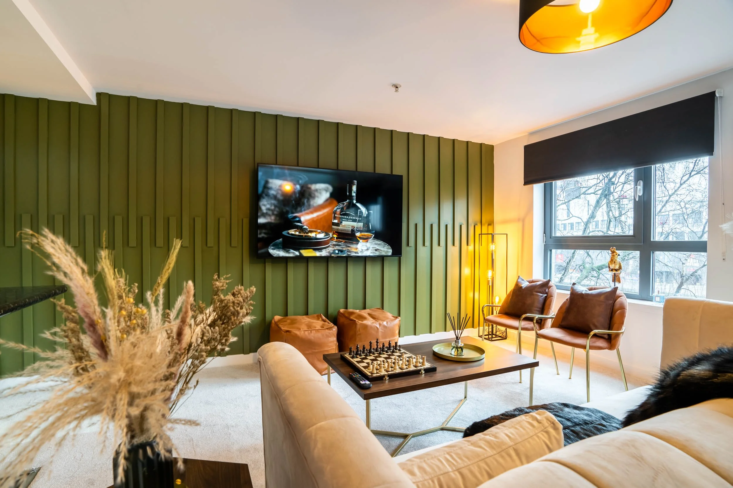 Living room with green accent wall, beige sofa, brown armchairs, a television, a coffee table with a chess set, and a large window with a view of trees outside.