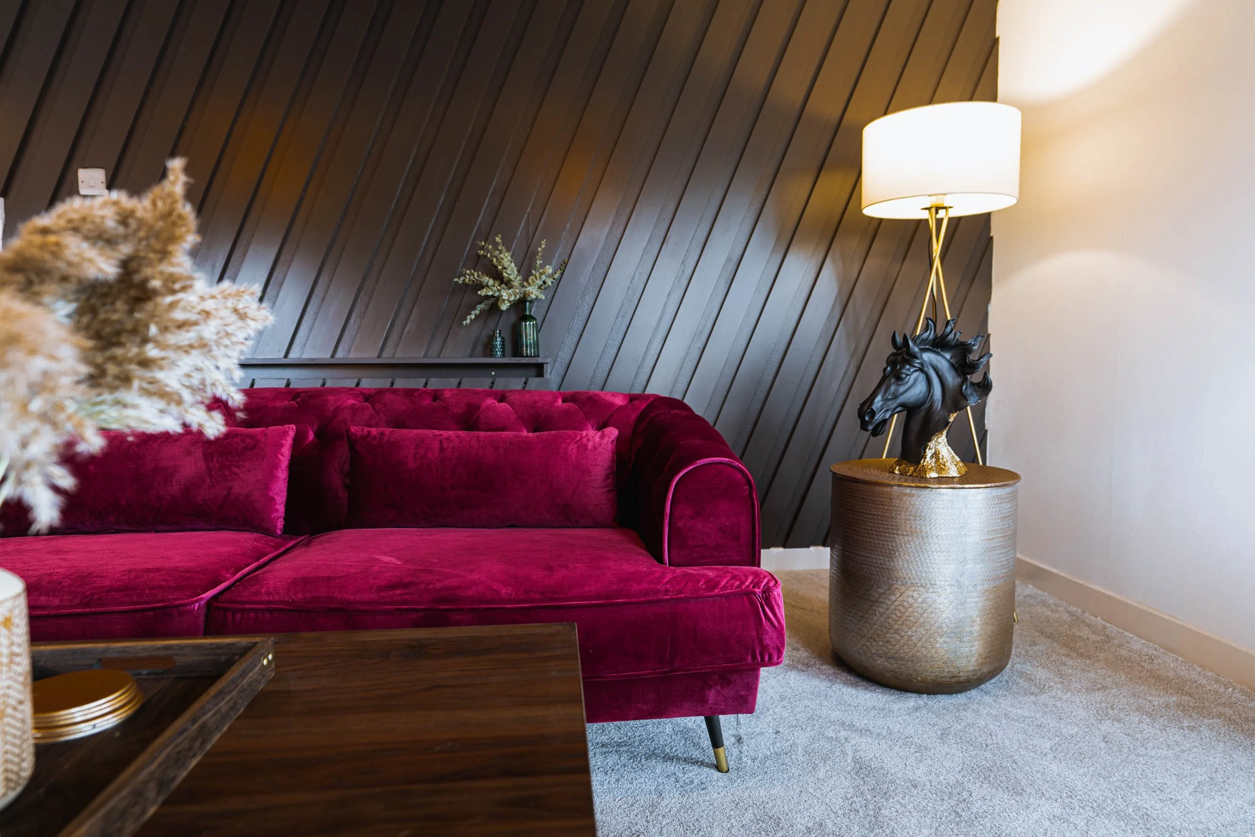 Living room with a deep pink velvet couch, a gold side table with a black horse head lamp, dark wood paneled wall, and a white floor lamp with a gold stand.