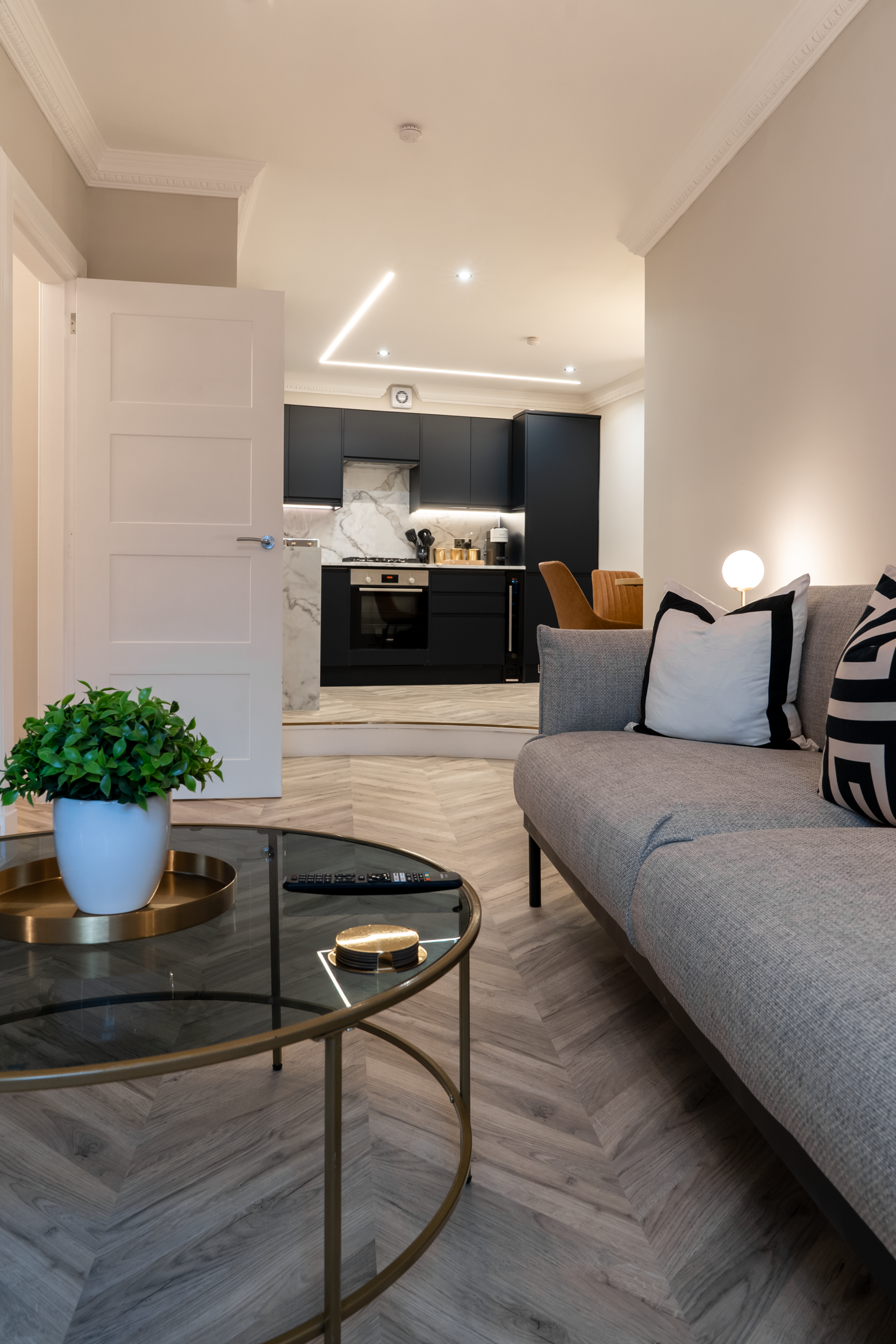 Living room with a gray sofa, black and white cushions, a round glass coffee table with a potted plant and remote control, and a kitchen with black cabinets, marble backsplash, and chrome appliances in the background.