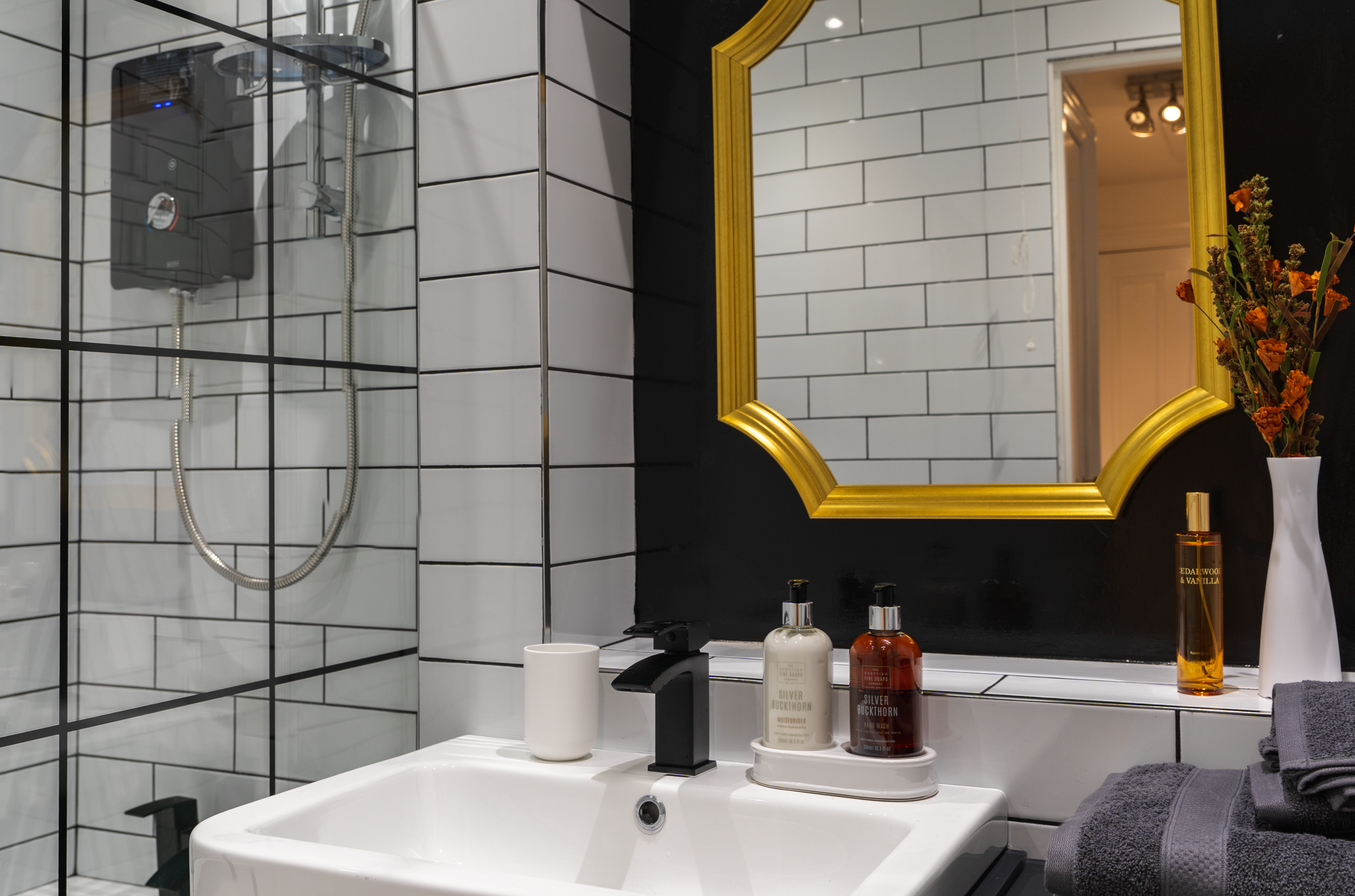 Bathroom sink with black faucet, white soap dispenser, amber and white bottles on a tray, a white cup, a tall white vase with orange and purple flowers, and a black towel, reflected in a large gold mirror, with a shower area with black and white tile