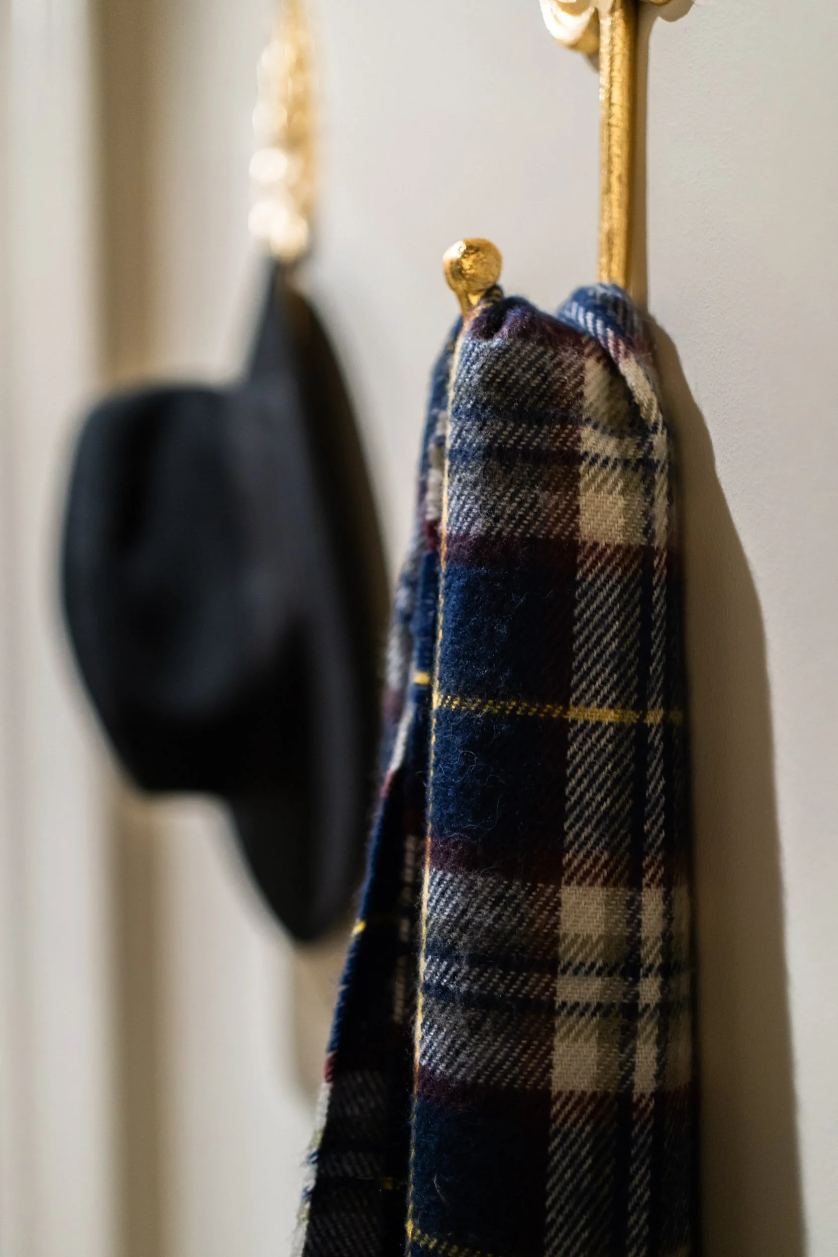 Close-up of a wall hook with a black hat and a plaid scarf hanging on it, against a beige wall.