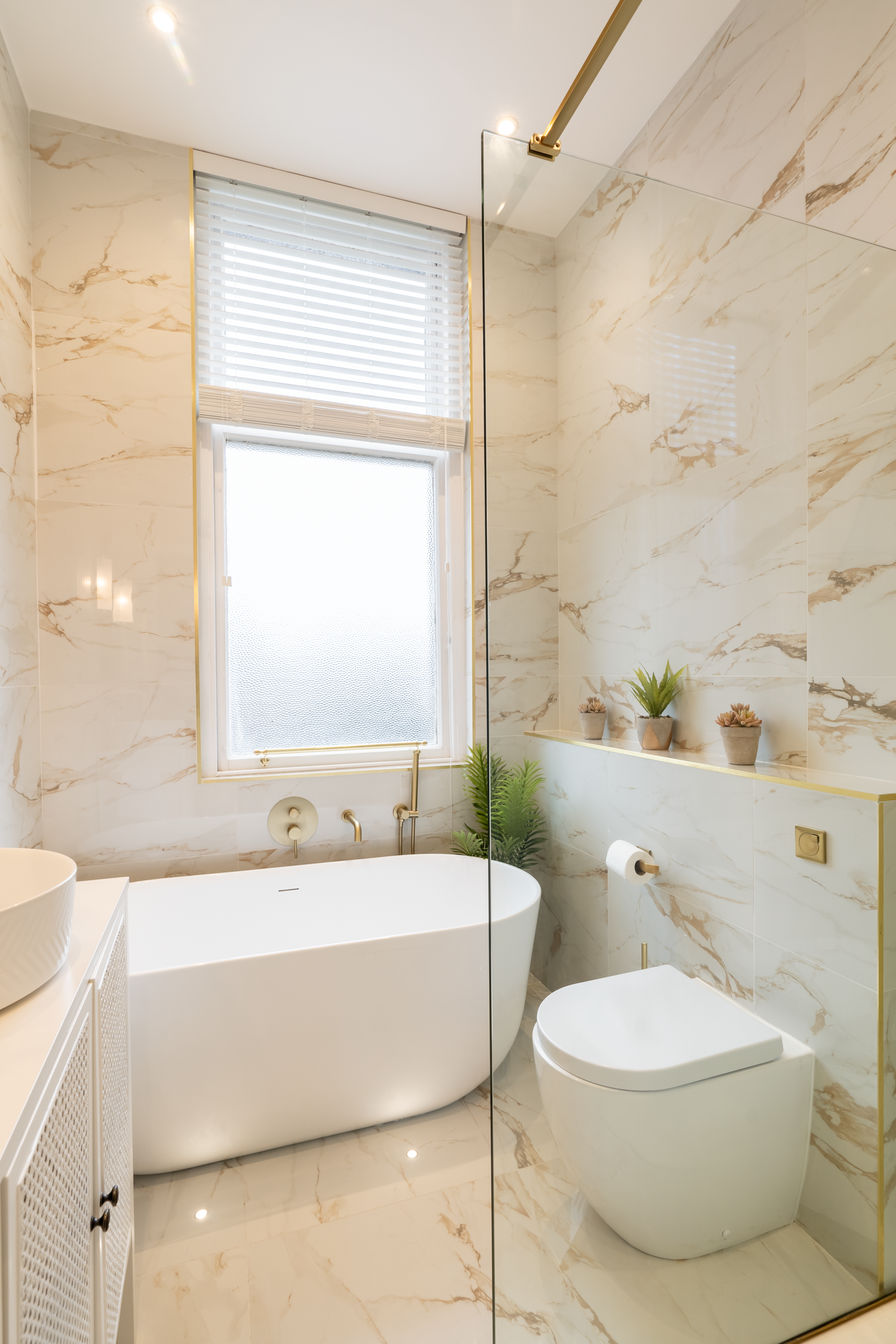 Modern bathroom with marble walls and floor, a freestanding bathtub under a window, a wall-mounted toilet, and decorative plants on a shelf.