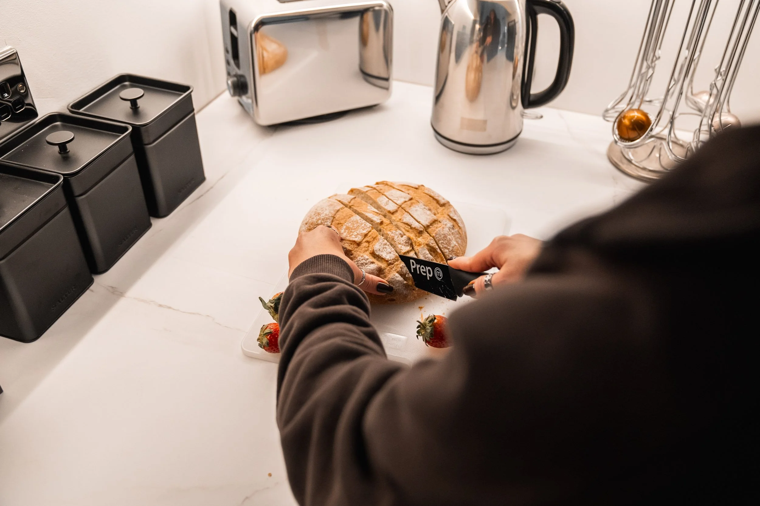 Person cutting a loaf of bread on a kitchen counter with a knife labeled 'Prep'. Strawberry fruits are also on the counter.