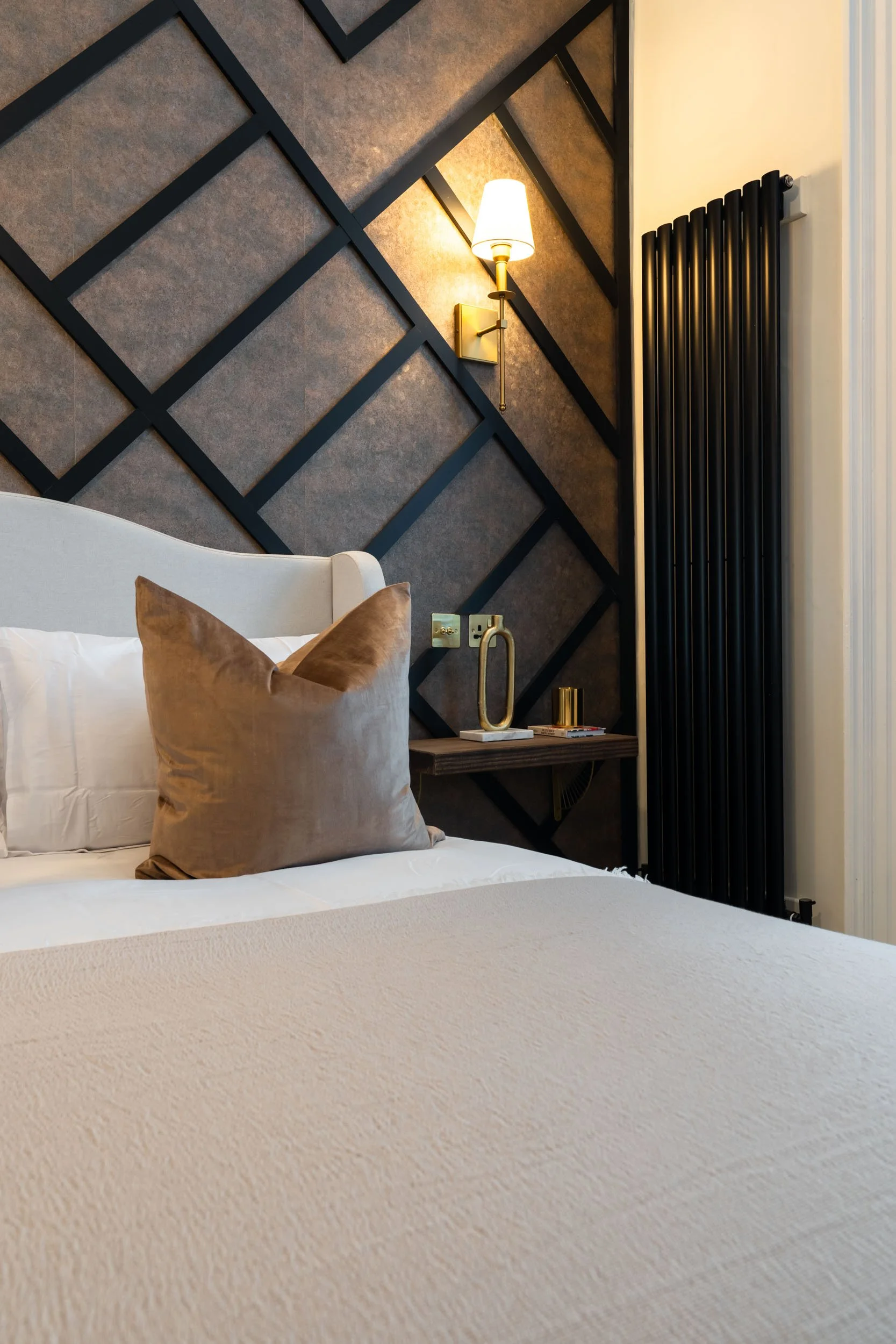 Close-up of a bedroom with a bed, decorative pillows, a geometric wall design, a wall-mounted lamp, and a radiator.