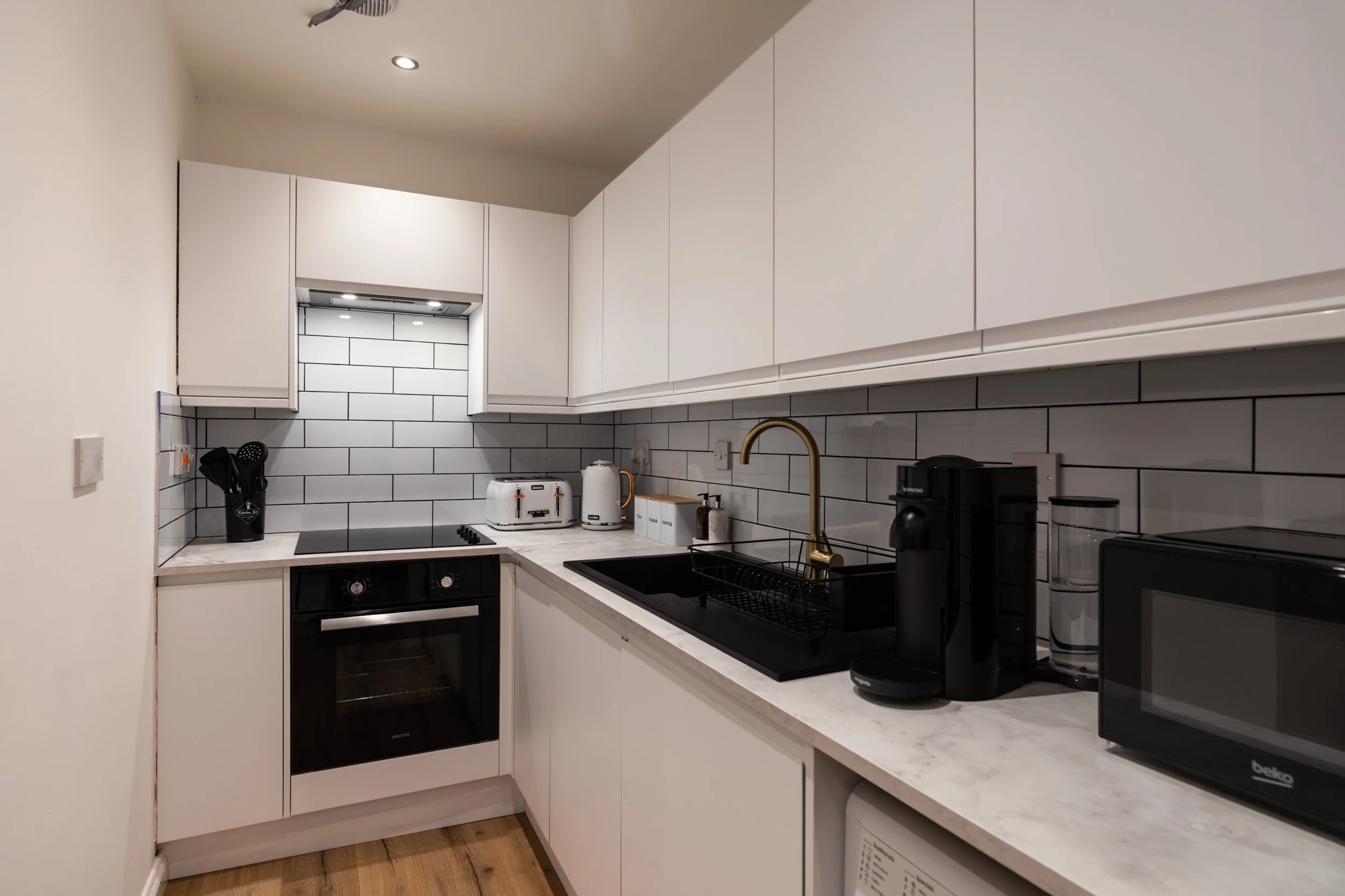Modern small kitchen with white cabinets, black and white backsplash, and various appliances including a toaster, kettle, coffee machine, microwave, and a black single oven with cooktop. Wood flooring.