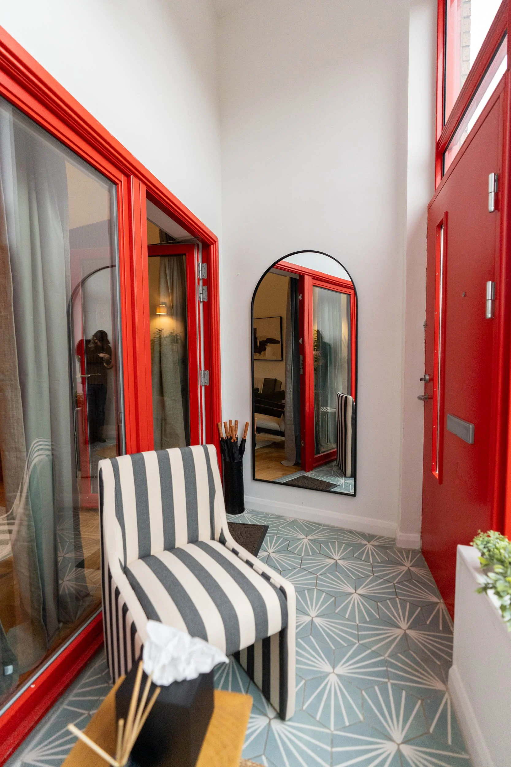 A cozy indoor porch with red-framed doors, a black and white striped armchair, a large mirror, and patterned tile flooring.