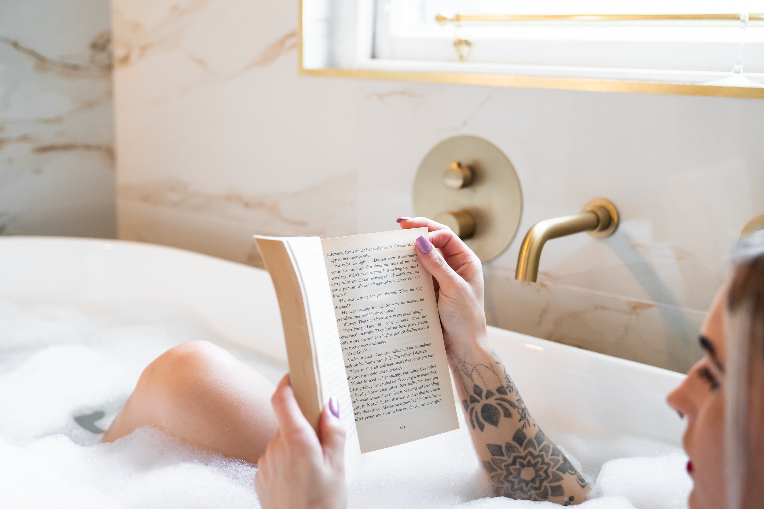 A person with a floral tattoo on their arm relaxing in a bubble bath, reading a book near a window with sunlight coming through.
