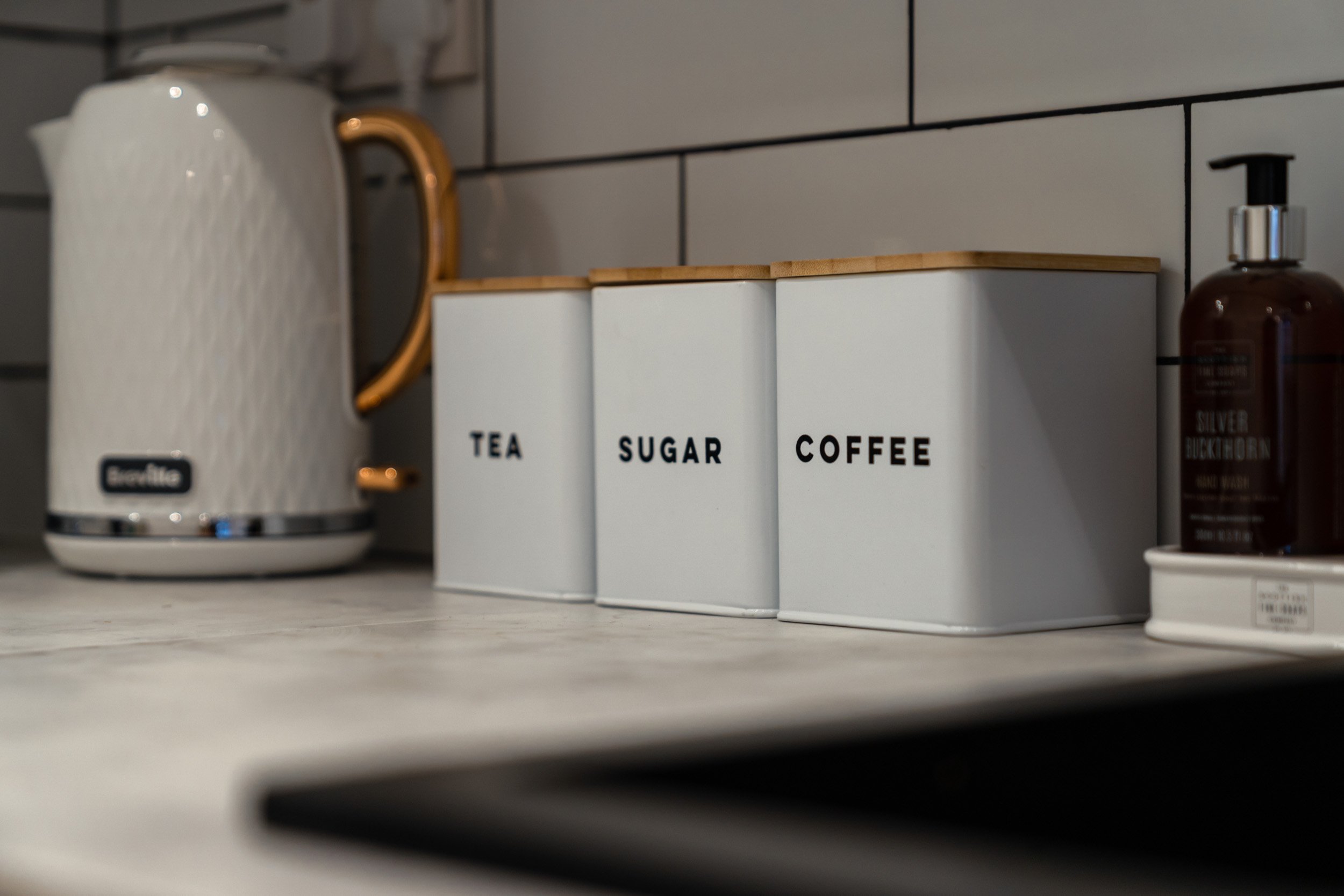 Kitchen countertop with a white textured electric kettle, white storage canisters labeled 'Tea', 'Sugar', and 'Coffee' with wooden lids, and a brown soap dispenser.