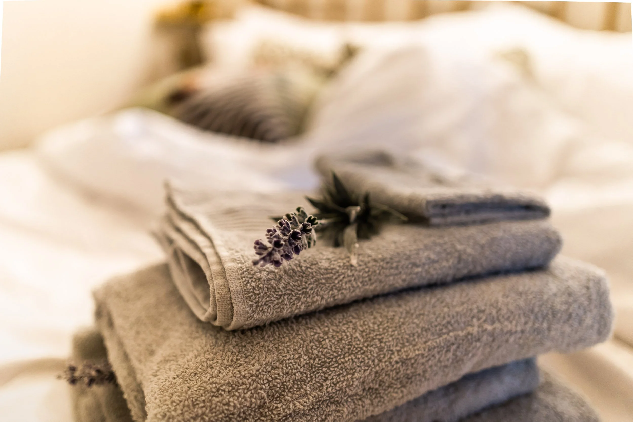 Stacked beige towels with a sprig of purple lavender on top, with a white shirt and a striped object in the background.