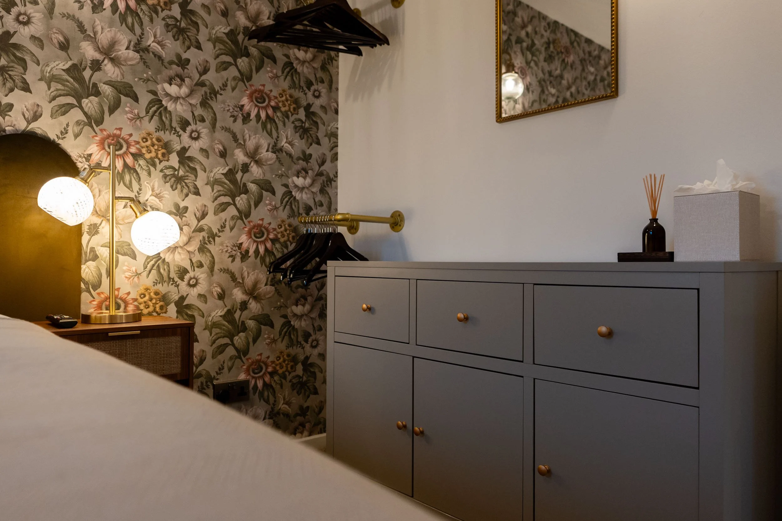 A bedroom corner with a floral wallpapered wall, a dresser with round gold knobs, a wall mirror with a gold frame, and a black vase with orange reed sticks and a tissue box on top. A yellow-lit table lamp with two white globe-shaped bulbs is on a sid