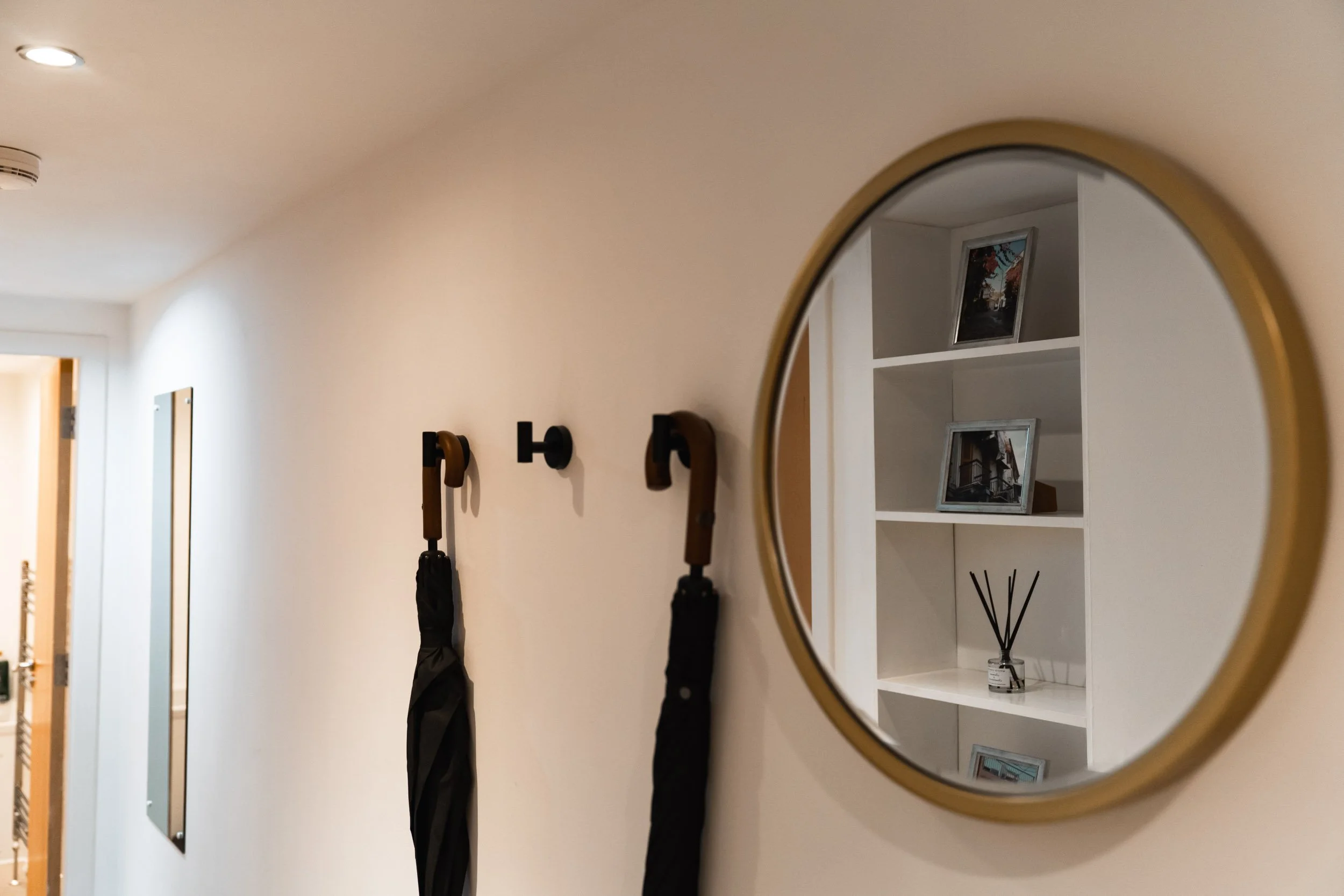 A hallway with a round mirror reflecting a white bookshelf with framed pictures and a reed diffuser. Two black hooks hold umbrellas on a beige wall.