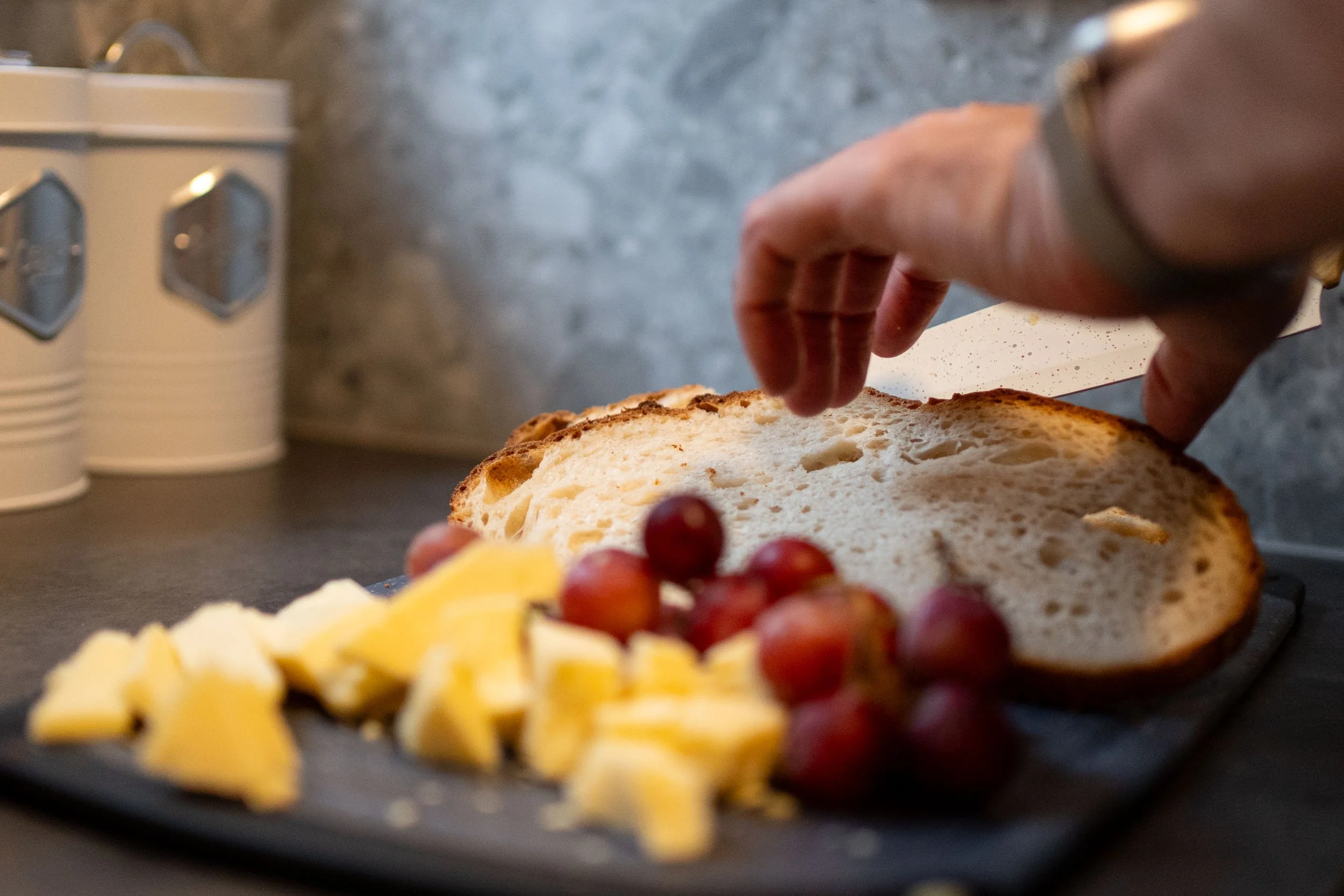 Person placing cheese and grapes on a slice of bread on a black cutting board in a kitchen.
