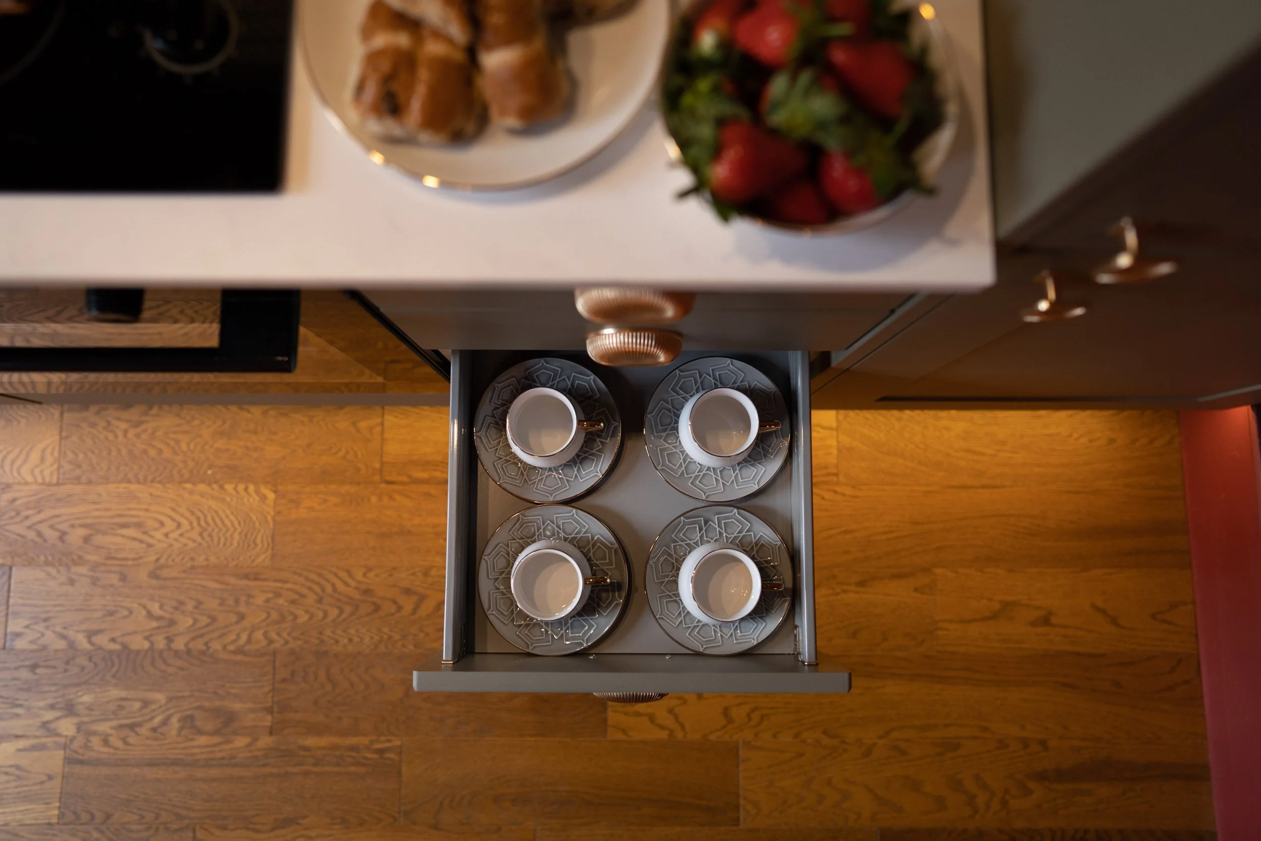 Four white coffee cups with matching saucers arranged in the drawer beneath a kitchen counter. The counter has bowls of strawberries and baked goods.