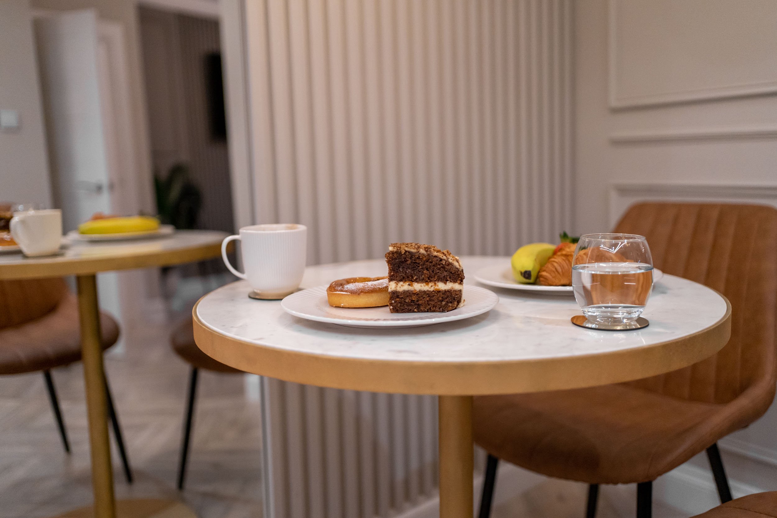 A round table with a slice of chocolate cake, a donut, a cup of coffee, a glass of water, a pear, and a croissant in a cozy dining area.