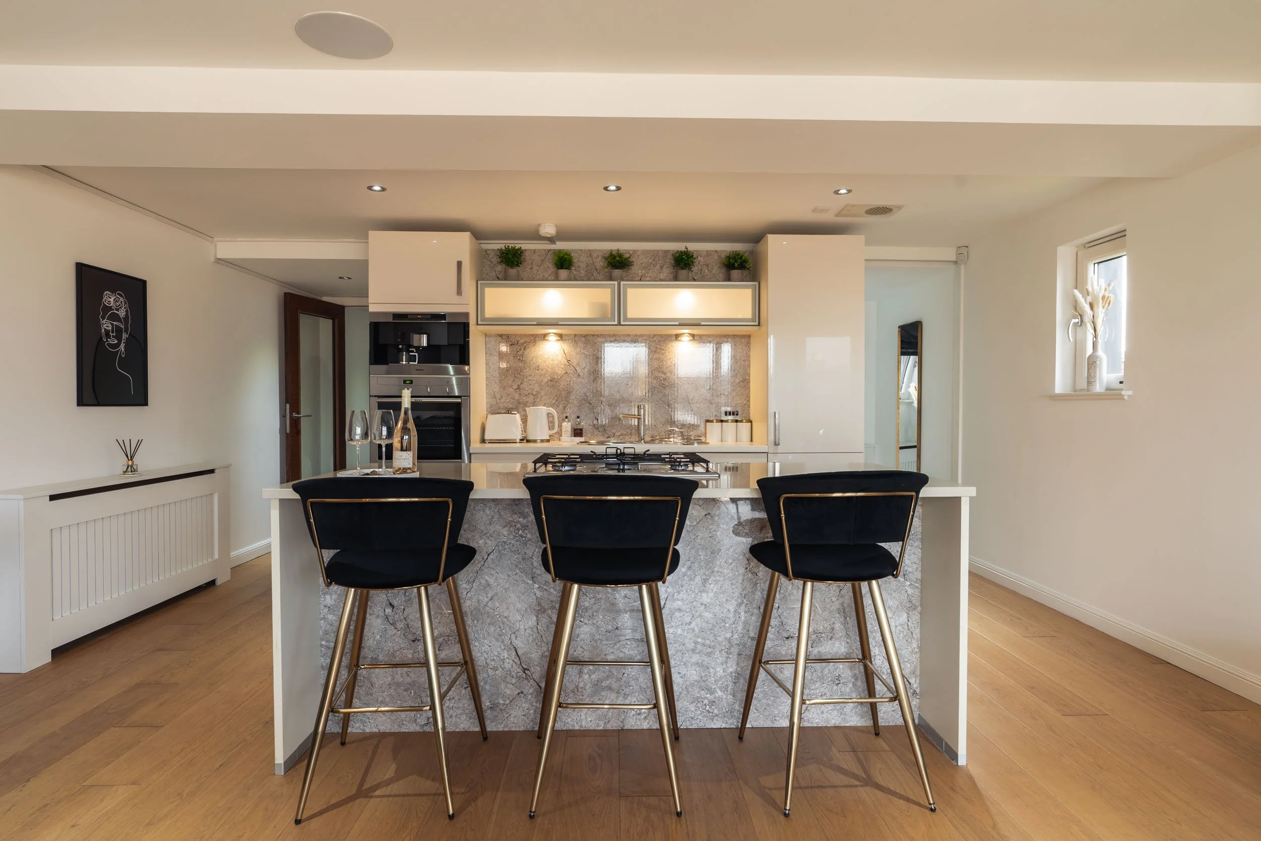 Modern kitchen with a marble island, black barstools with gold legs, white cabinetry, appliances, and decorative plants on top.