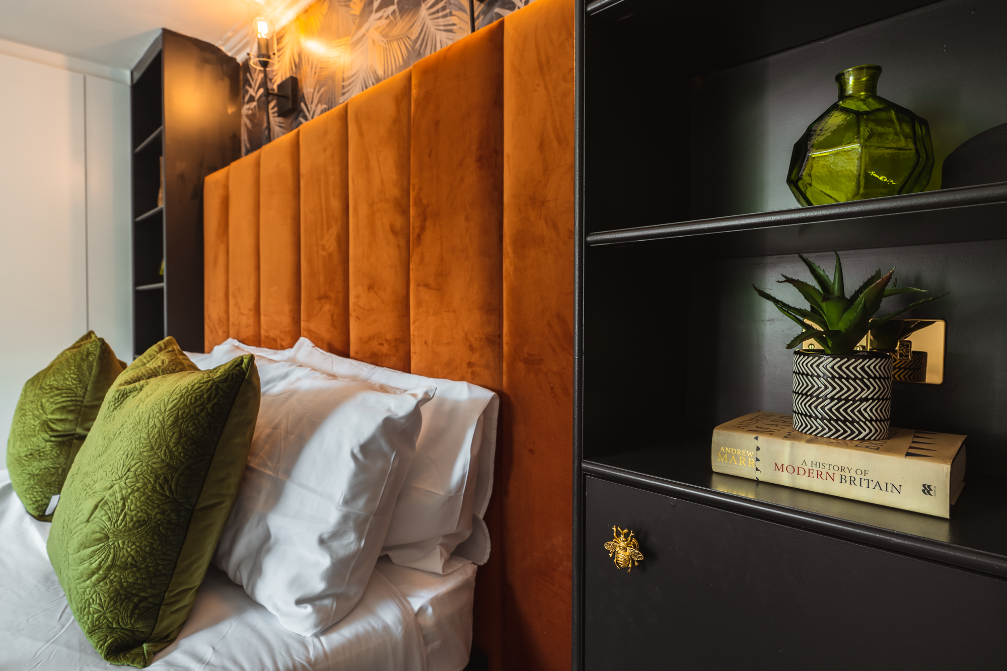 A bedroom with a bed that has white sheets, green textured pillows, and an orange headboard. There is a black bookshelf next to the bed containing a green vase, a potted plant, and books, including one titled "A History of Modern Britain."