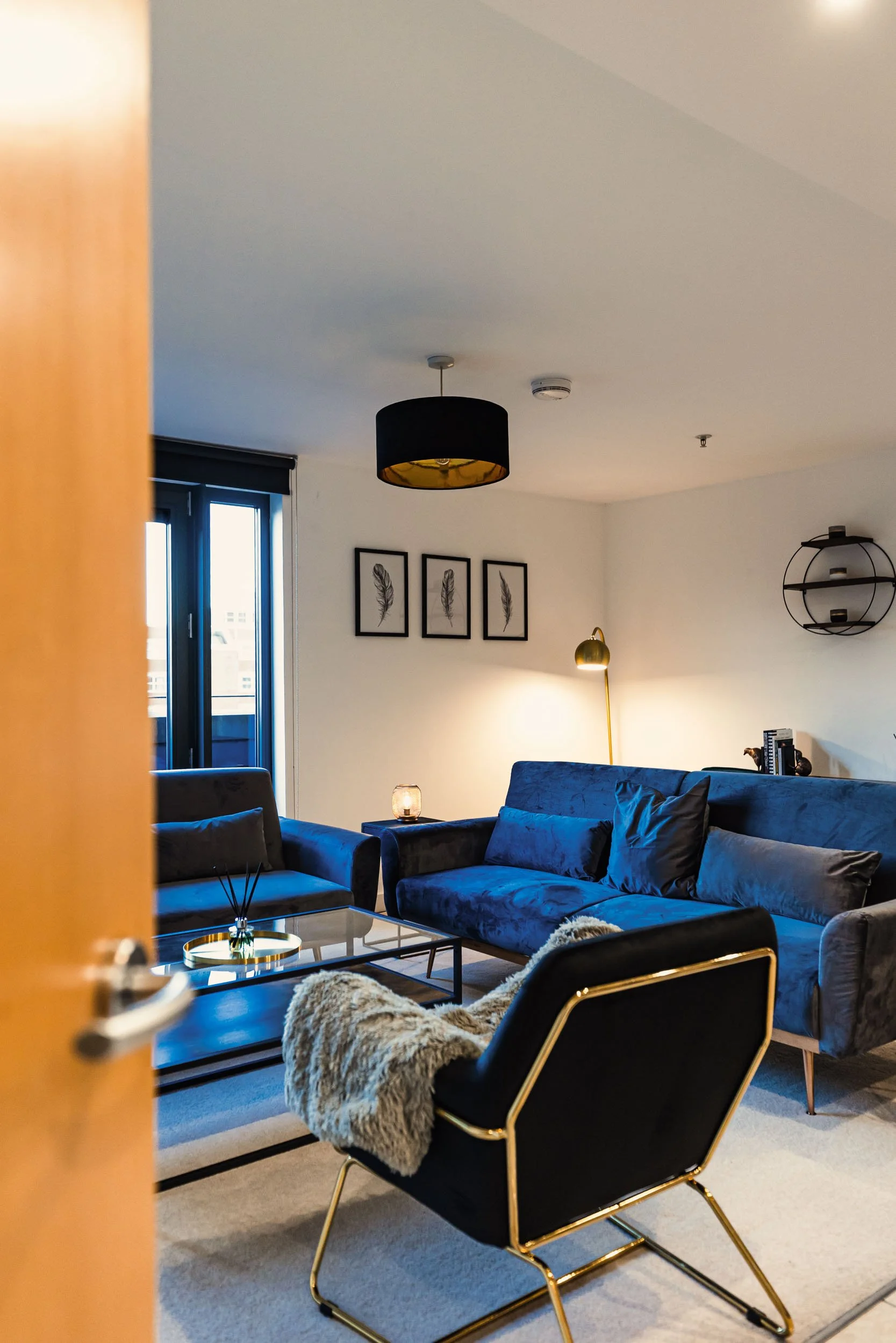 Modern living room with blue velvet sofas, black armchair with gold frame, glass coffee table, wall art of feathers, standing gold lamp, round black ceiling light, and balcony door