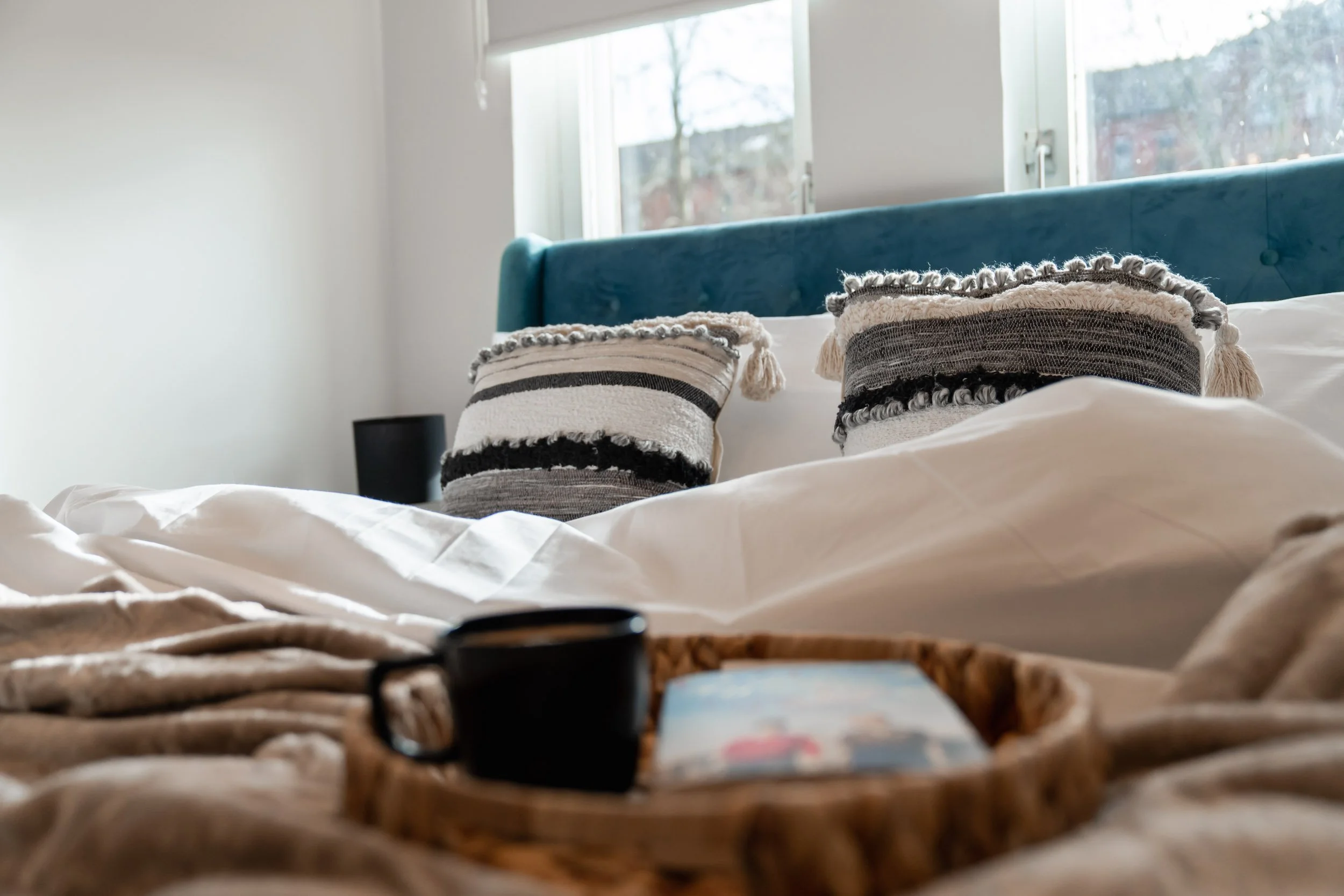 A bed with two decorative pillows, a teal headboard, and a tray with a coffee mug and a book in the foreground, in a bright bedroom with large windows.