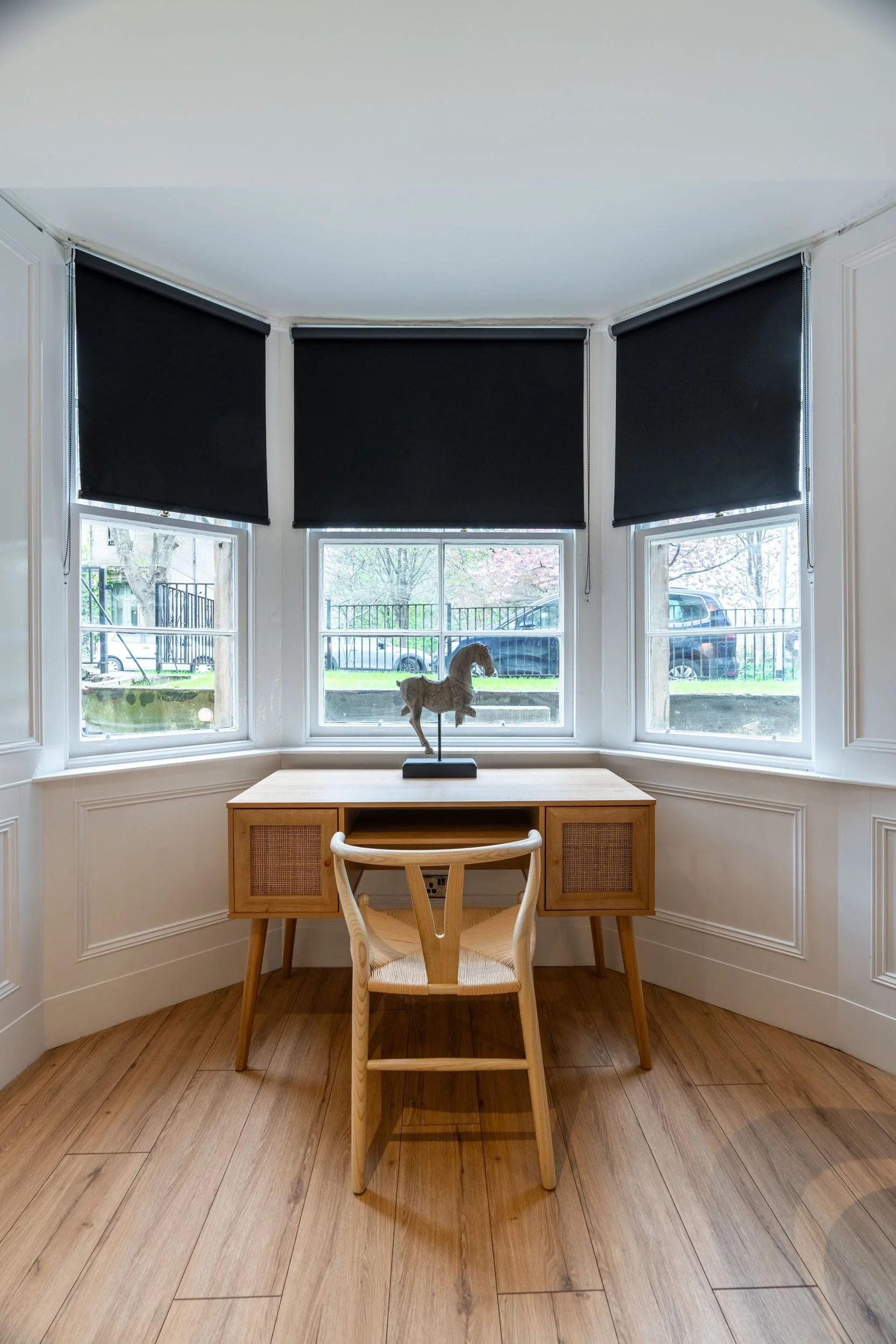 A small wooden desk with a beige chair in front of a bay window with three sections. The window has black window shades. On the desk is a decorative horse figurine on a stand.