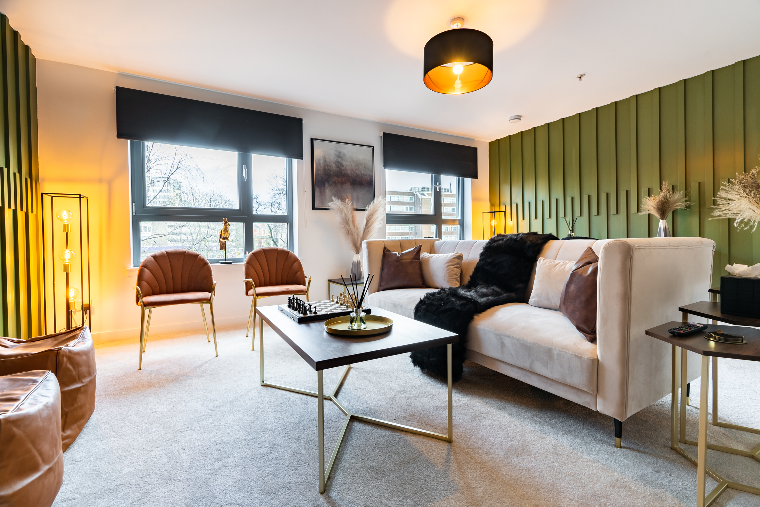 Modern living room with a white sofa, brown and beige cushions, a black fur throw, a coffee table with chess and a tray, two brown chairs, side tables, decorative plants, lamps, a large window, and a green accent wall.