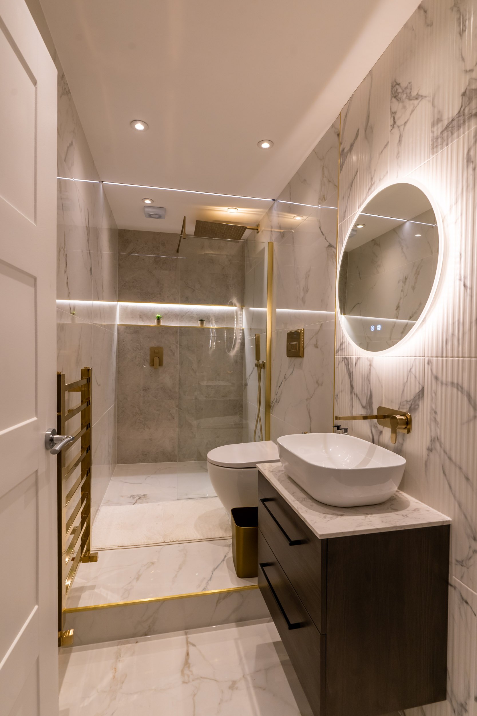 Modern bathroom with marble walls and floors, a vessel sink on a dark vanity, a round backlit mirror, a toilet, a walk-in shower with glass enclosure, gold fixtures, and recessed ceiling lights.