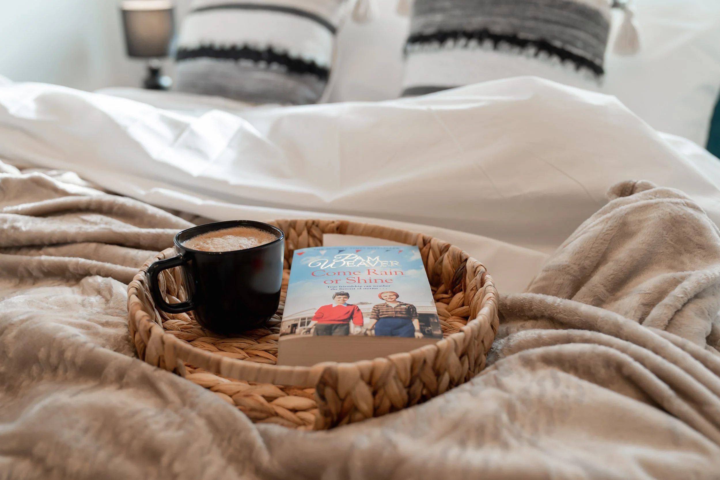 A wicker tray with a black coffee mug and a book titled 'I Am Grace, Come Rain or Shine' sitting on beige and cream-colored bedding in a softly lit bedroom.