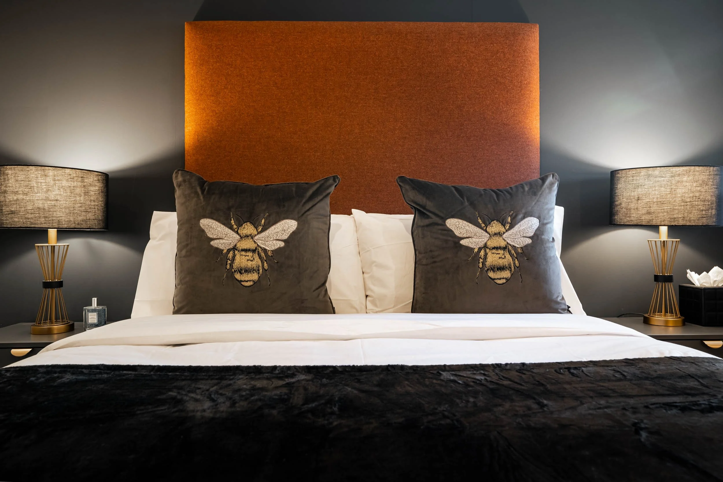 A neatly made bed with two decorative pillows featuring a bee embroidery, flanked by two table lamps on either side with warm light, in a modern bedroom with a large upholstered headboard and dark wall background.