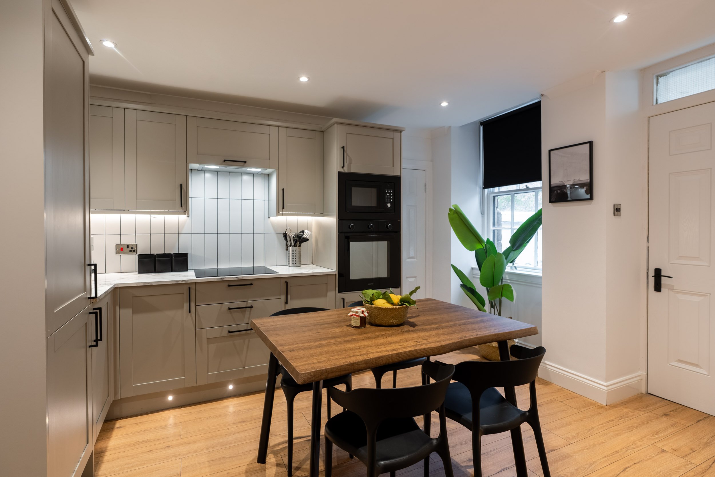 Modern kitchen with beige cabinets, white tiled backsplash, black appliances, a wooden dining table, black chairs, and a large window with a black roller blind, decorated with a green plant.