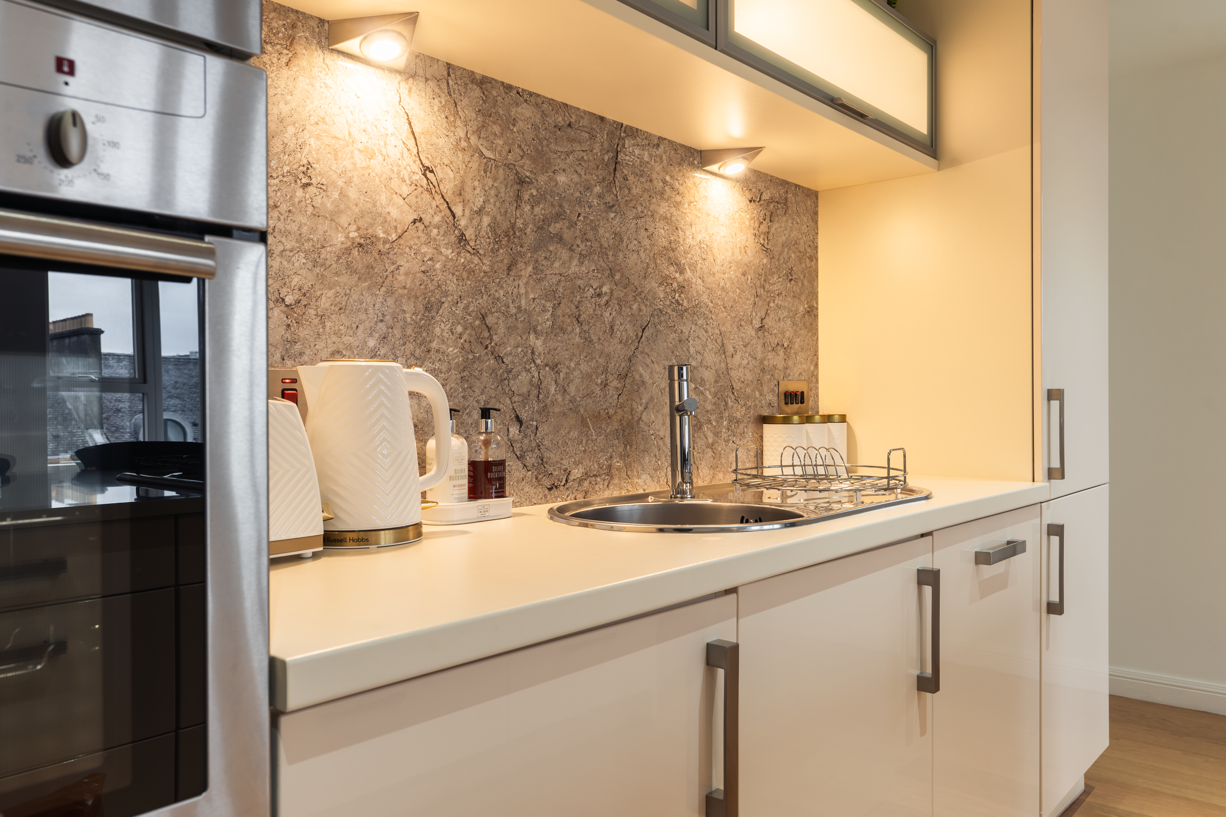 Modern kitchen with beige cabinets, marble backsplash, small stainless steel sink, kettle, soap dispenser, toaster, and countertop on a light-colored surface.