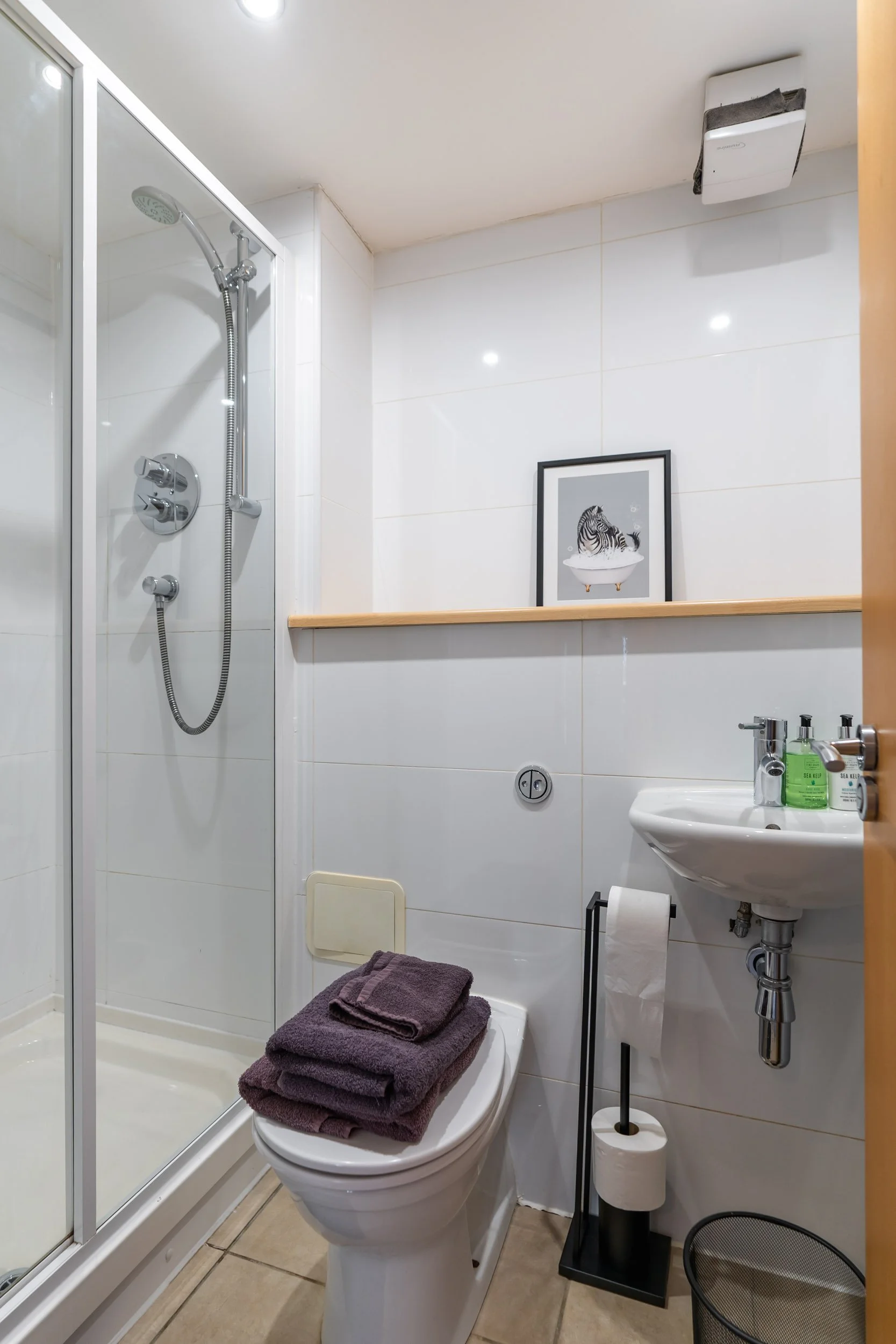Small bathroom with a shower enclosure, a toilet with folded towels on top, a wall-mounted sink with green soap dispensers, a framed zebra art print, a toilet paper holder, and a trash bin.
