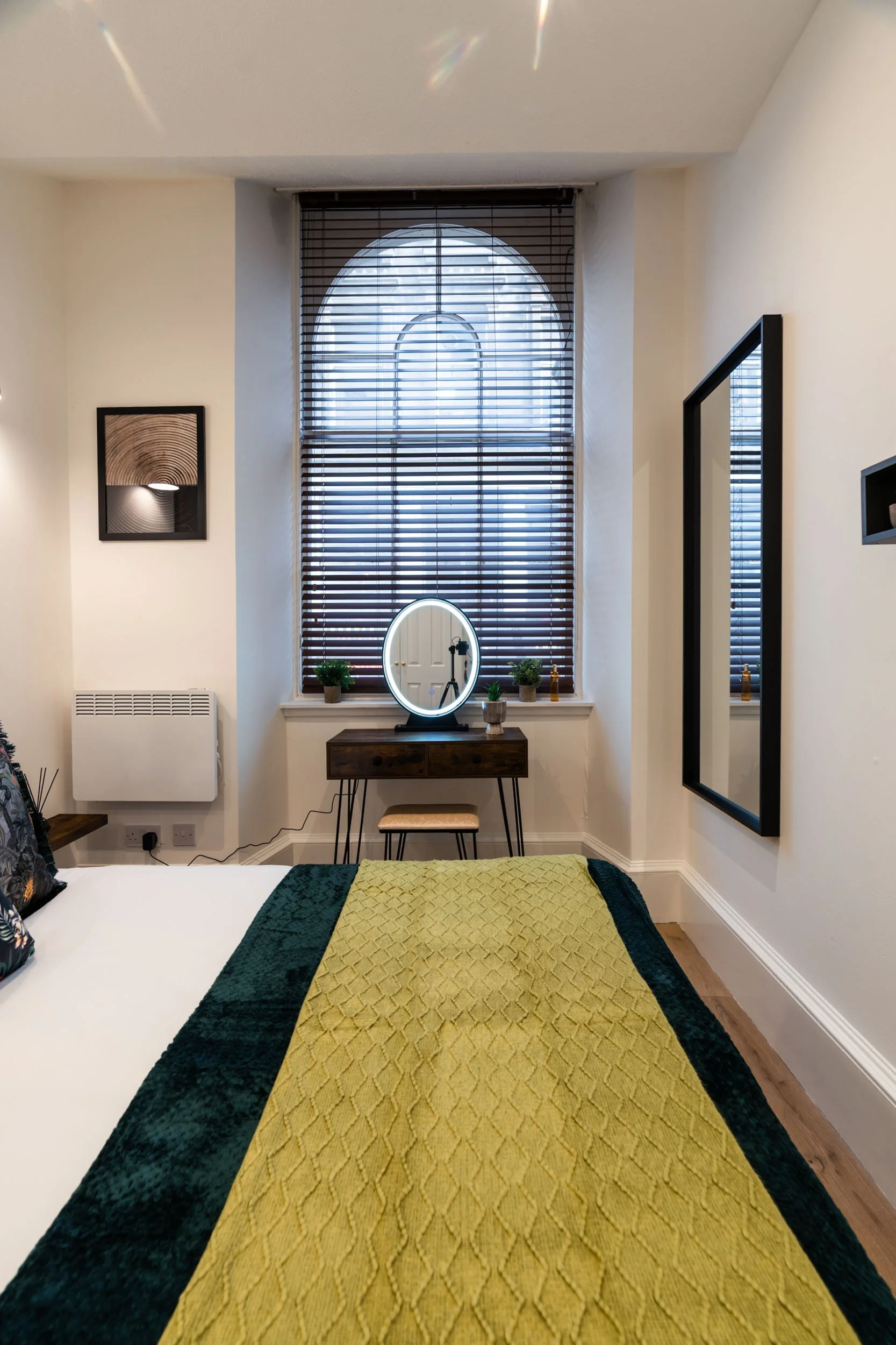 A bedroom with a large arched window with wooden blinds, a vanity with a round illuminated mirror, small potted plants, and a stool. Part of a bed with a yellow and green quilt and decorative pillows is visible in the foreground.