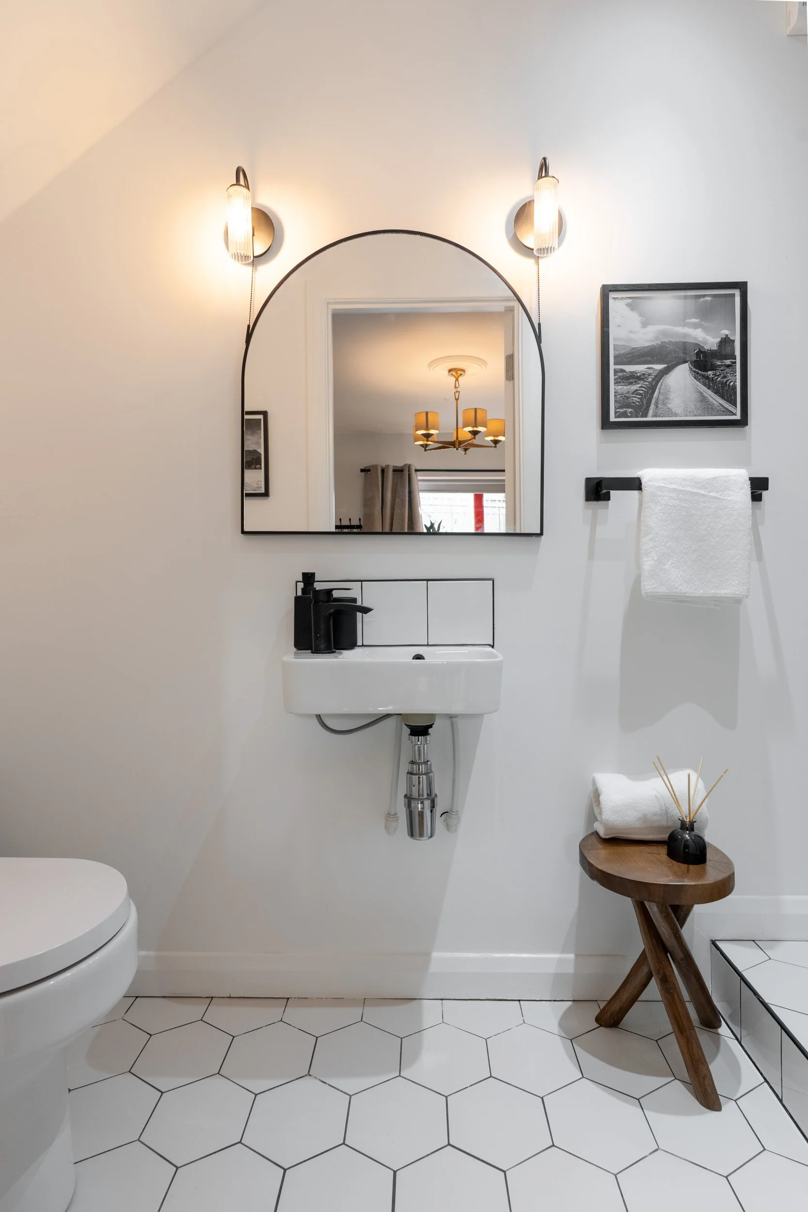 A modern bathroom with a small white sink, a large arched mirror, black wall sconces, a black picture frame, a white towel on a black towel rack, a small wooden stool with a rolled towel and reed diffuser, and white hexagonal floor tiles.