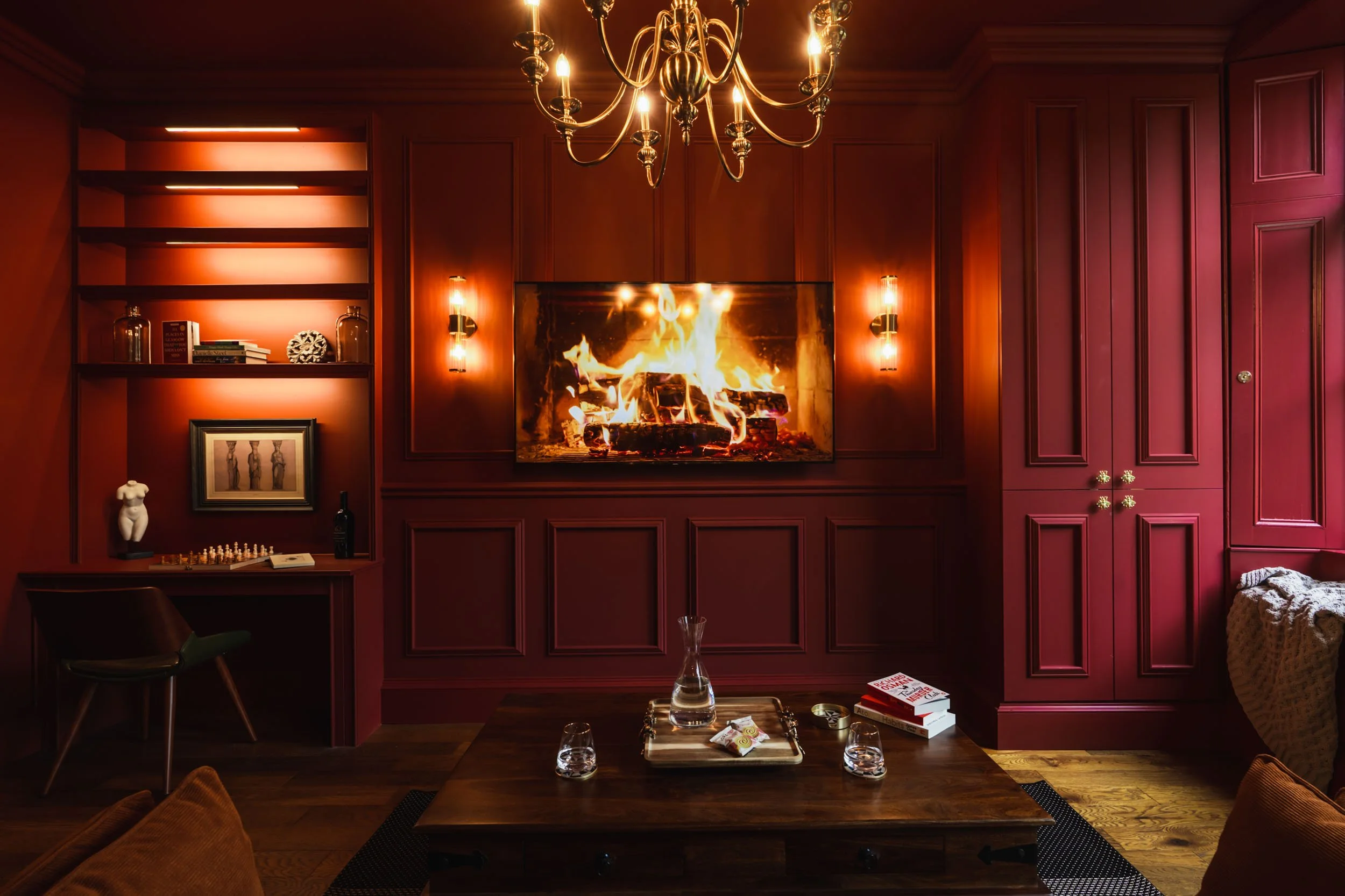 Living room with a fireplace, mounted TV showing a fire, dark red wood paneling, chandelier, bookshelf, small desk with a chess set, and a coffee table with books and glasses.