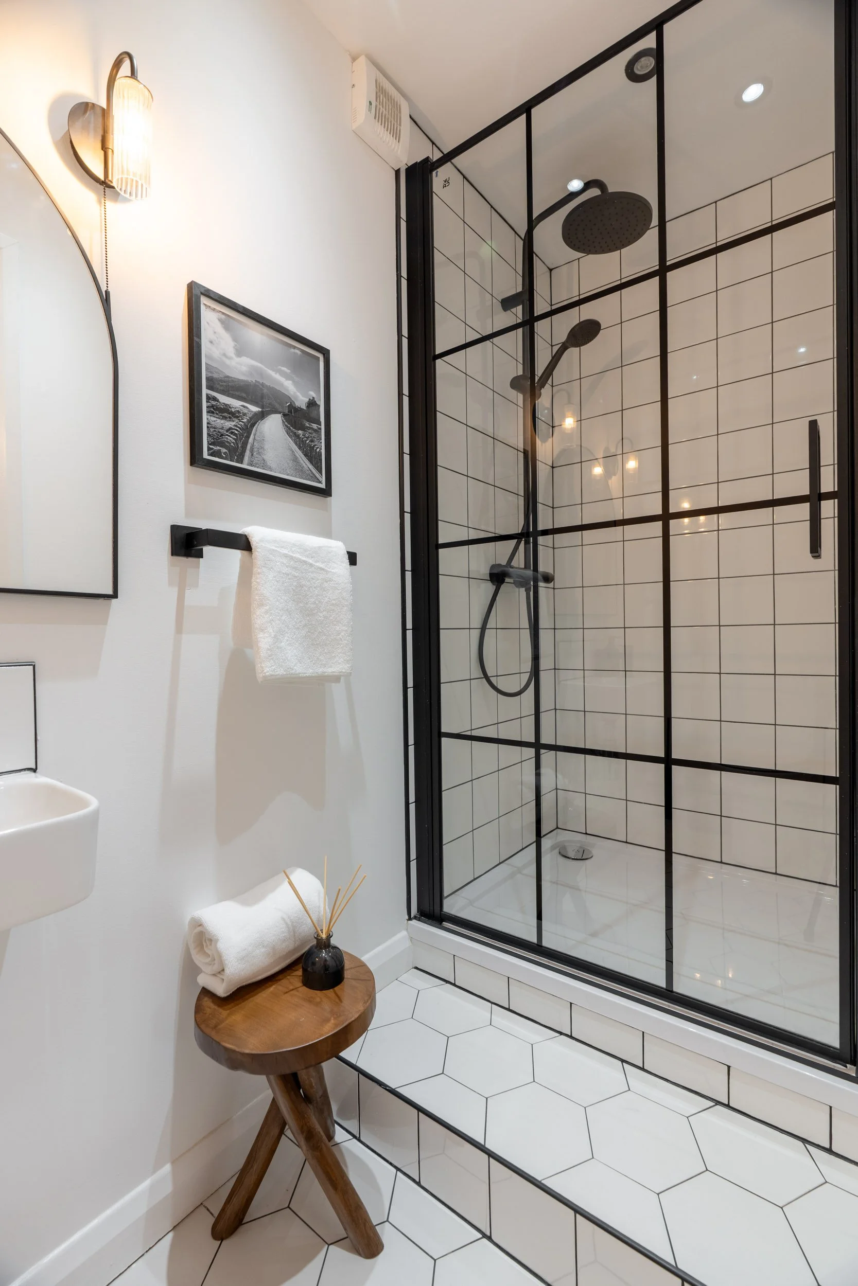 Modern bathroom with a glass enclosed shower, black hardware, white hexagonal floor tiles, a small wooden stool with rolled white towels and a black reed diffuser, a white wall-mounted sink, a black and white landscape photograph, a wall-mounted scon