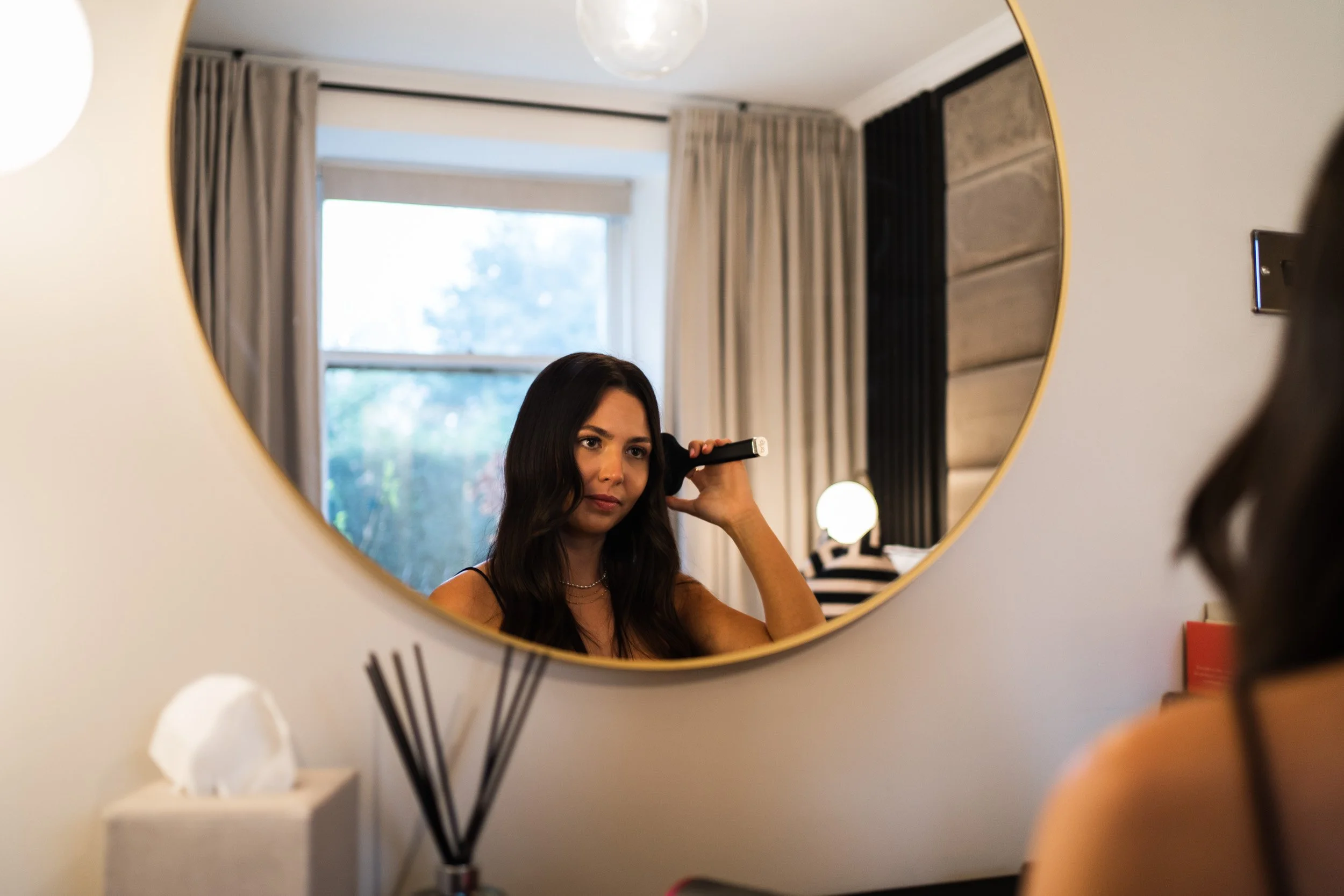 A woman with long dark hair looking into a round mirror while holding a curling iron near her hair in a well-lit room with curtains and a window in the background.