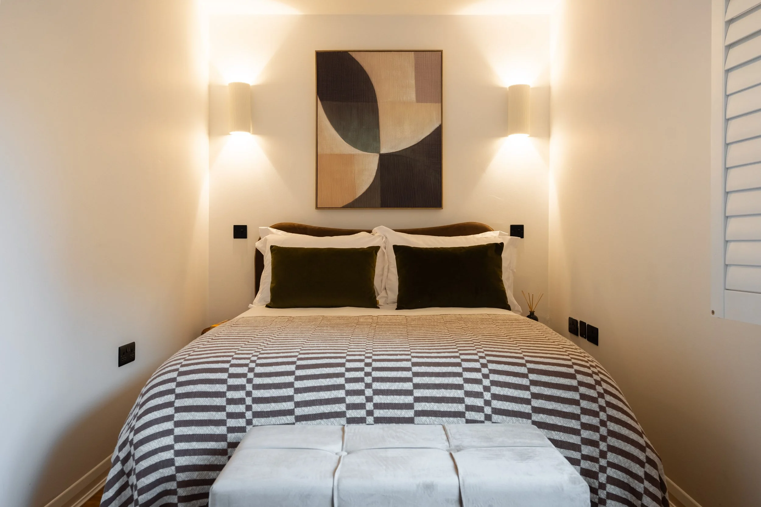 A neatly made bed with dark green pillows, a patterned bedspread, and a white ottoman at the foot, in a bedroom with minimalistic decor, wall sconces, abstract artwork, and light-colored walls.